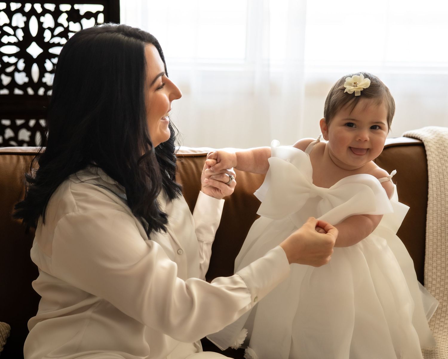 A dark-haired mother in white smiles warmly at her baby girl in a white dress on a leather chair.