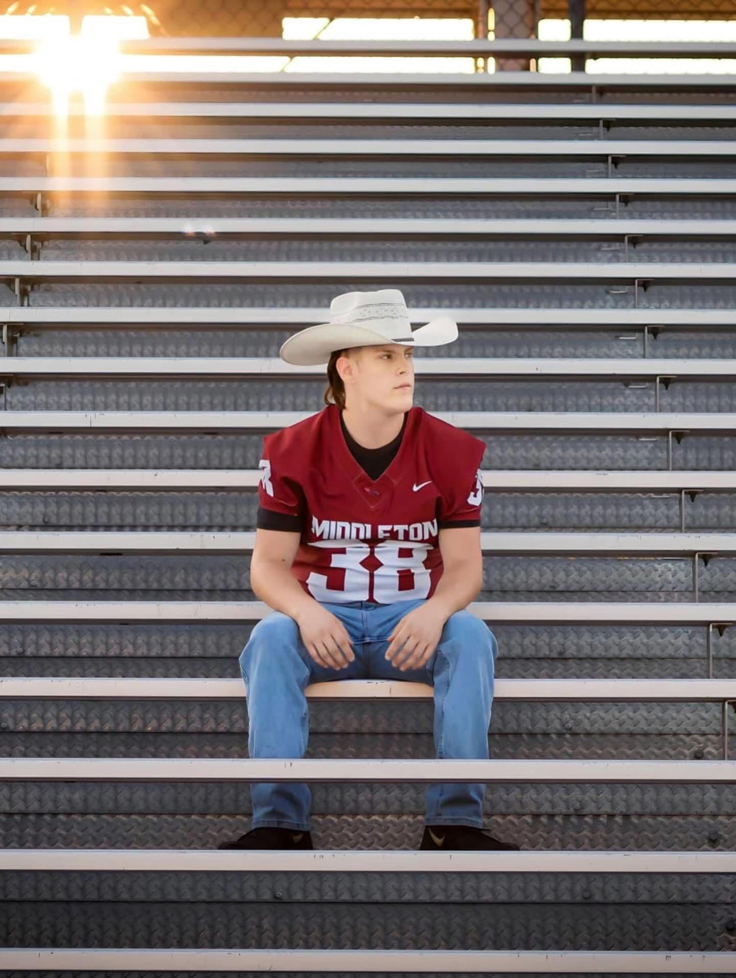 Teen boy in Middleton football jersey number 38 and cowboy hat sitting on stadium bleachers at sunset.