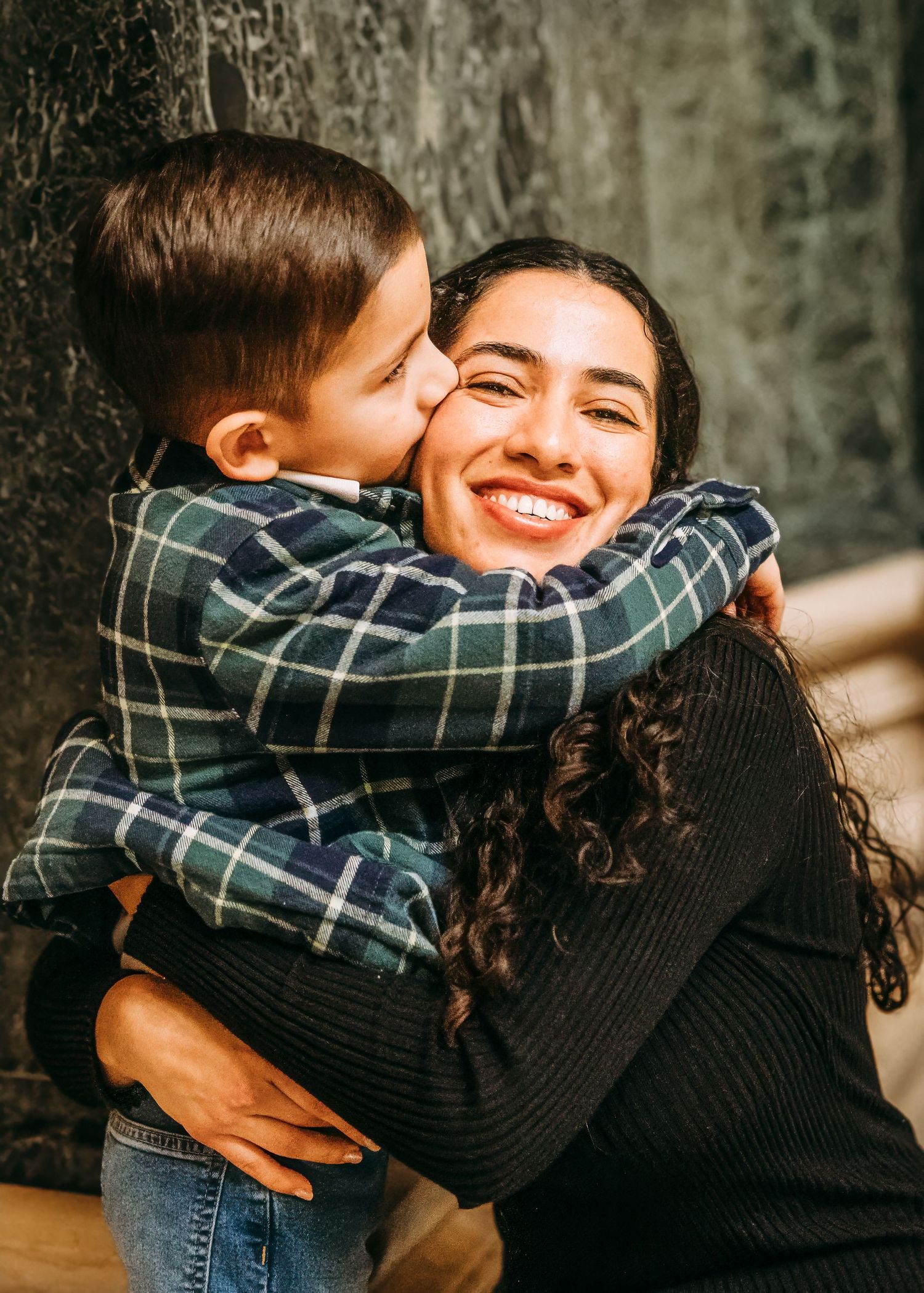 A smiling woman hugs a young boy in a plaid shirt who kisses her cheek.