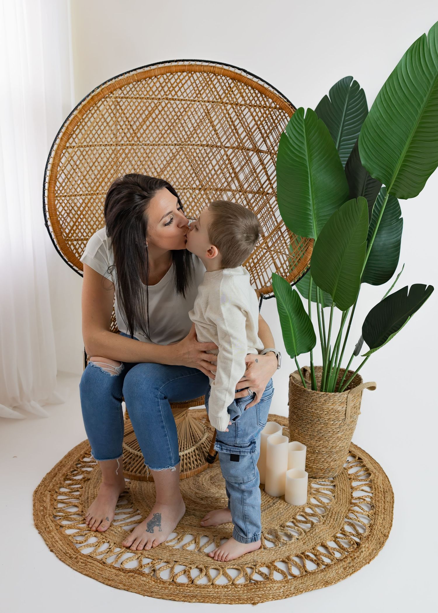 Dark-haired mother kissing young son beside rattan peacock chair and tropical plant indoors.