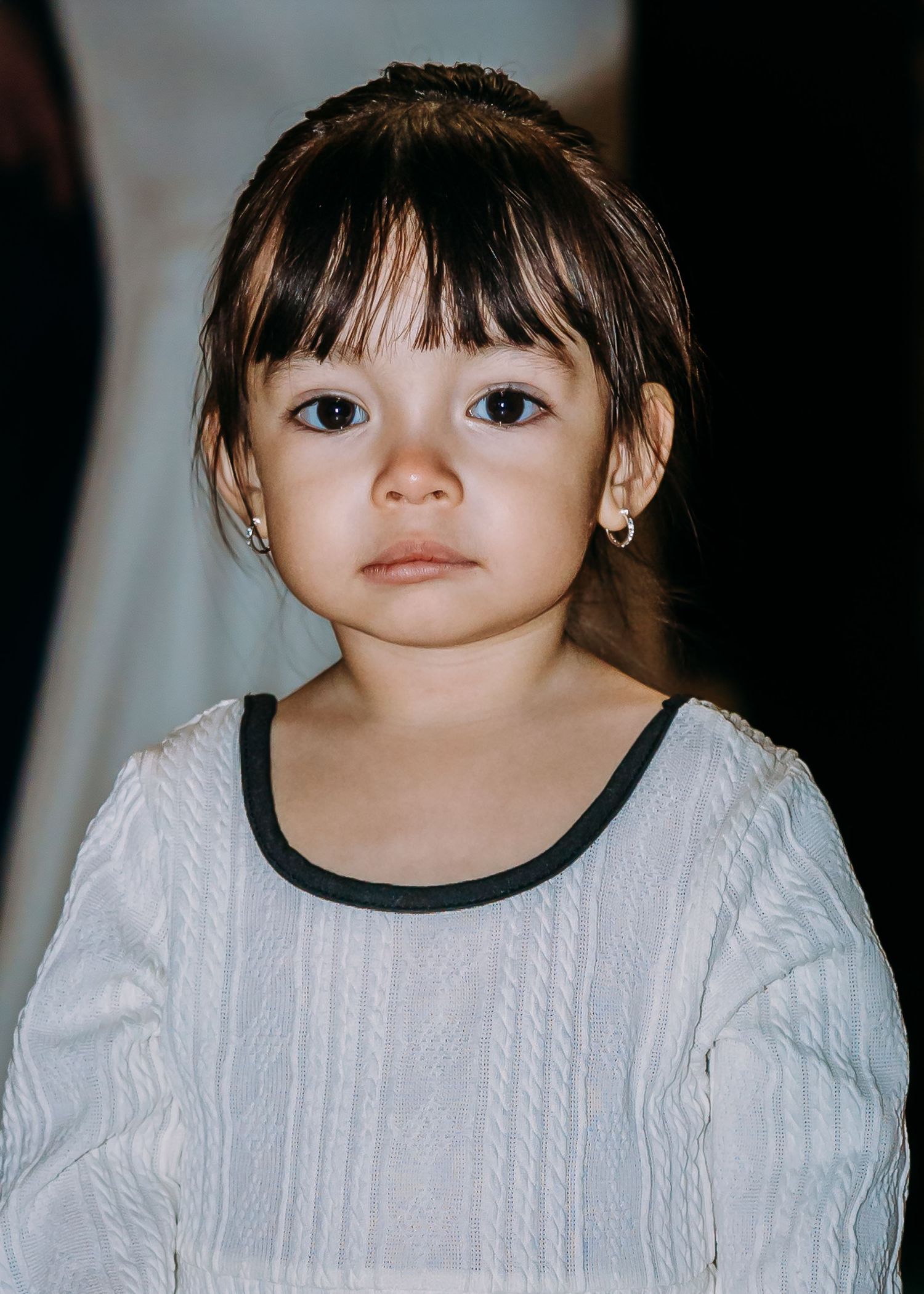 Close-up portrait of a young girl with bangs, wearing a white top with black trim and small hoop earrings.