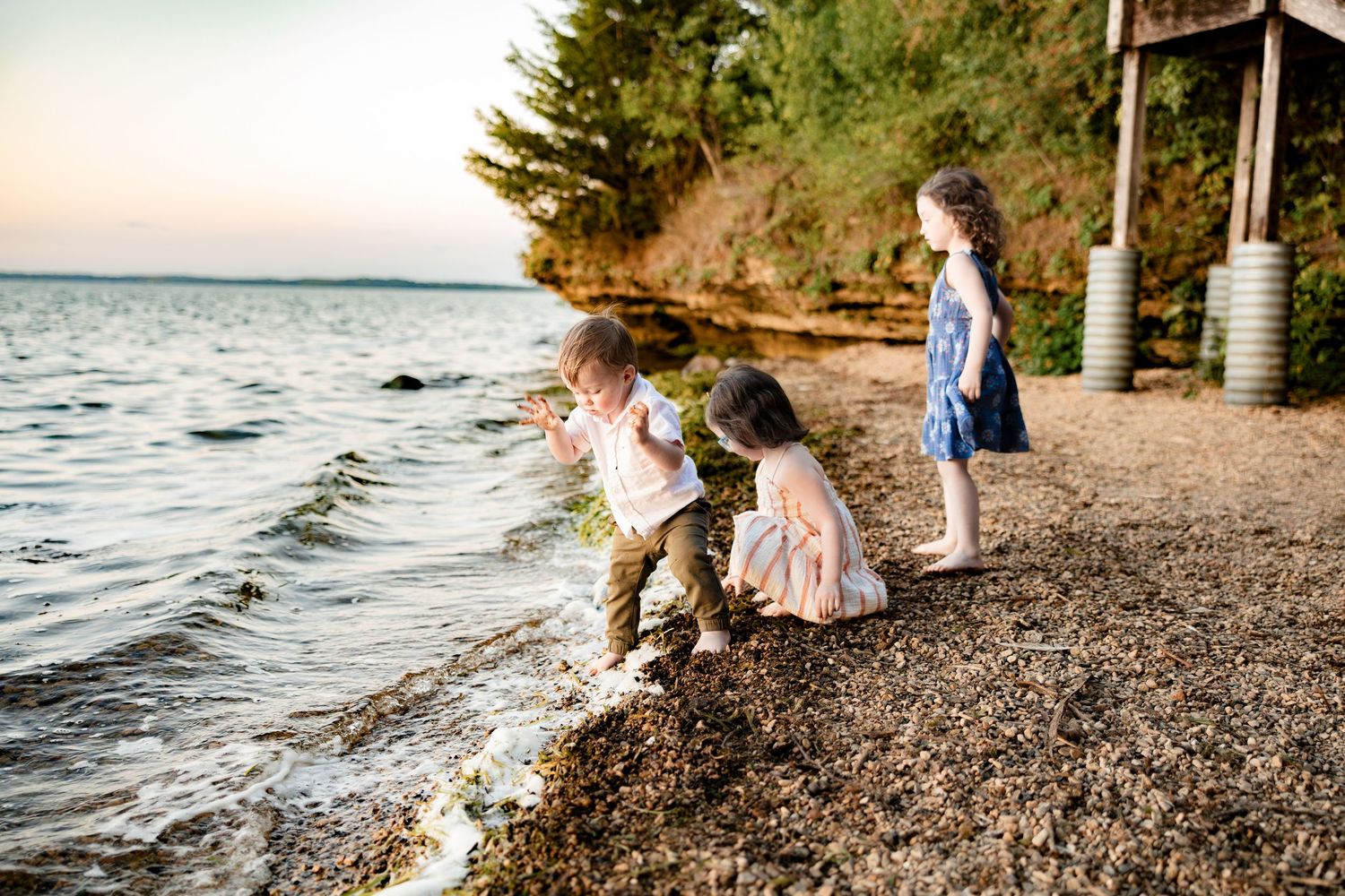 Three young children playing on a rocky shoreline at sunset, with calm water and trees in the background.