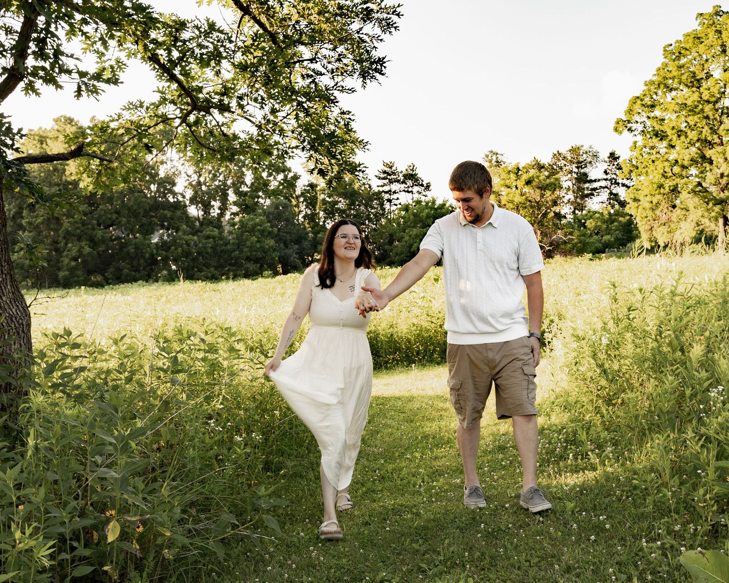 A couple walks hand-in-hand through a lush green park, smiling at each other on a sunny summer day.