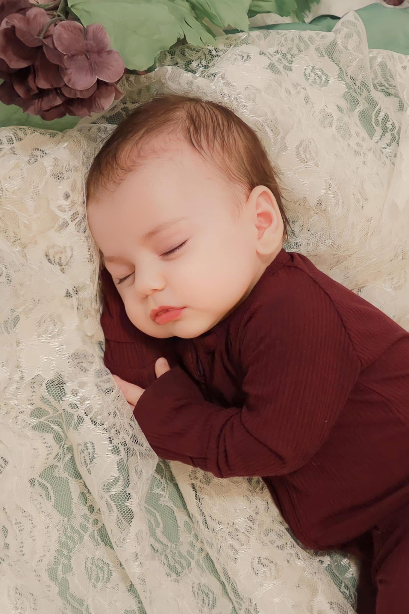 A peaceful newborn baby in a burgundy onesie sleeps on delicate white lace with floral decor.