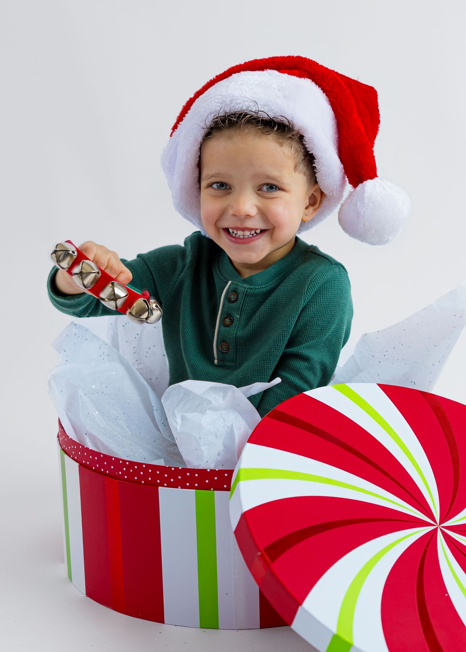 Happy child in Santa hat sitting inside large gift box holding jingle bells for Christmas.