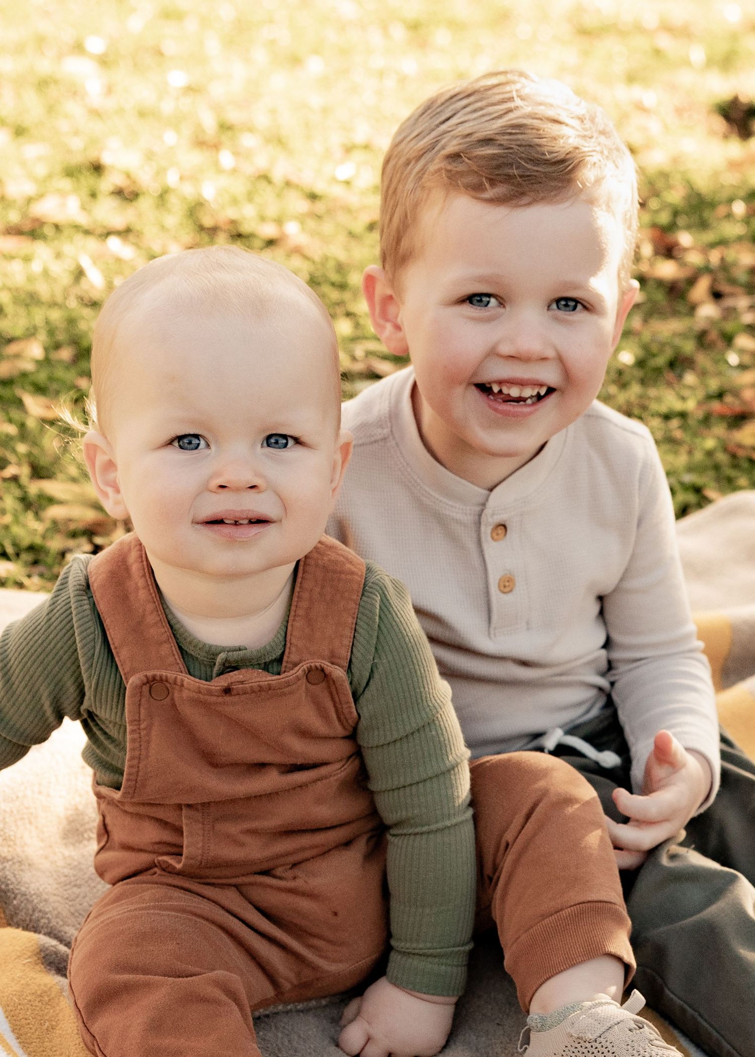 Two young brothers smile warmly while sitting together on a blanket in a sunny autumn park.