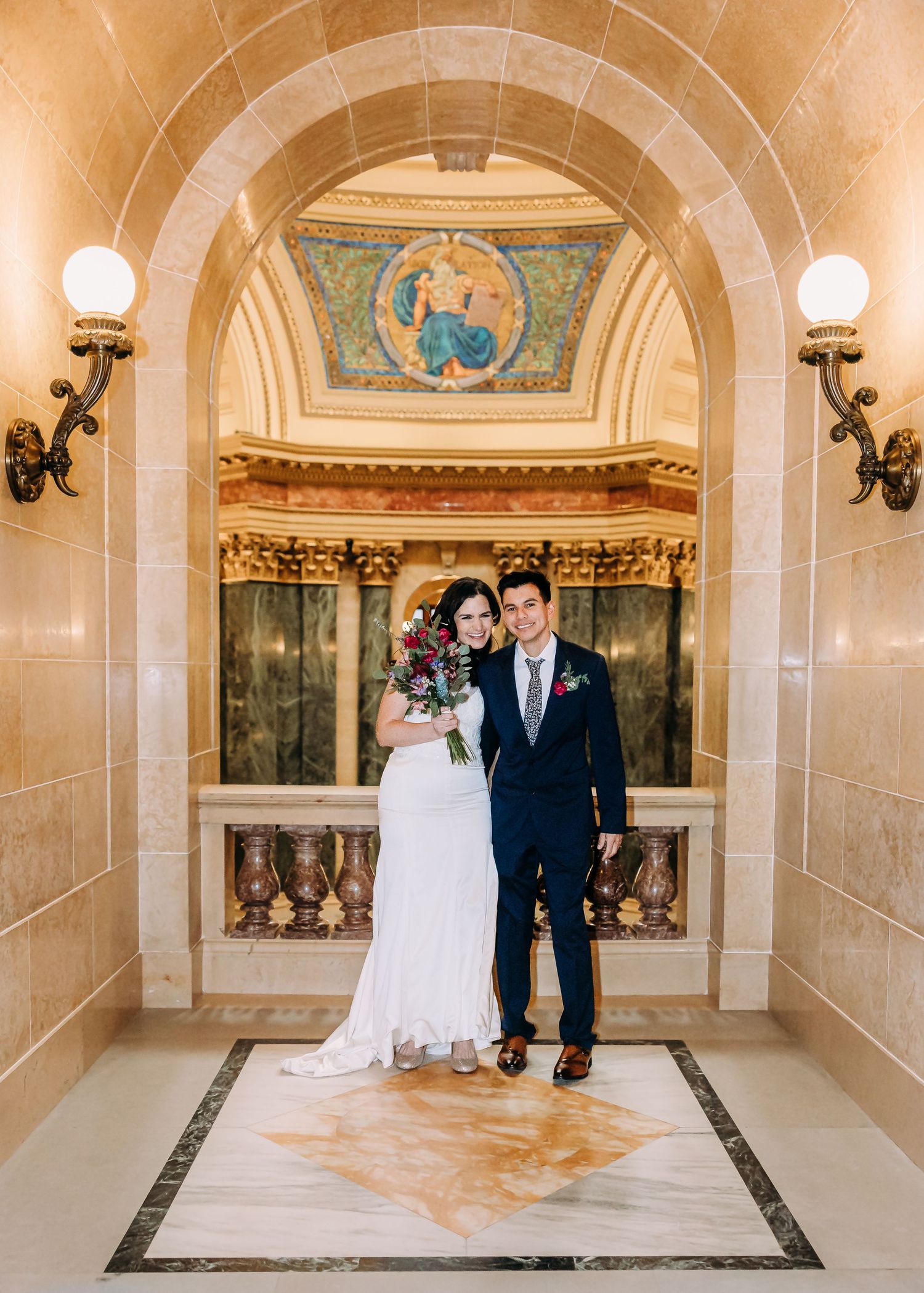 Bride with red bouquet and groom in navy suit posing under ornate arch with mosaic in grand historic venue.