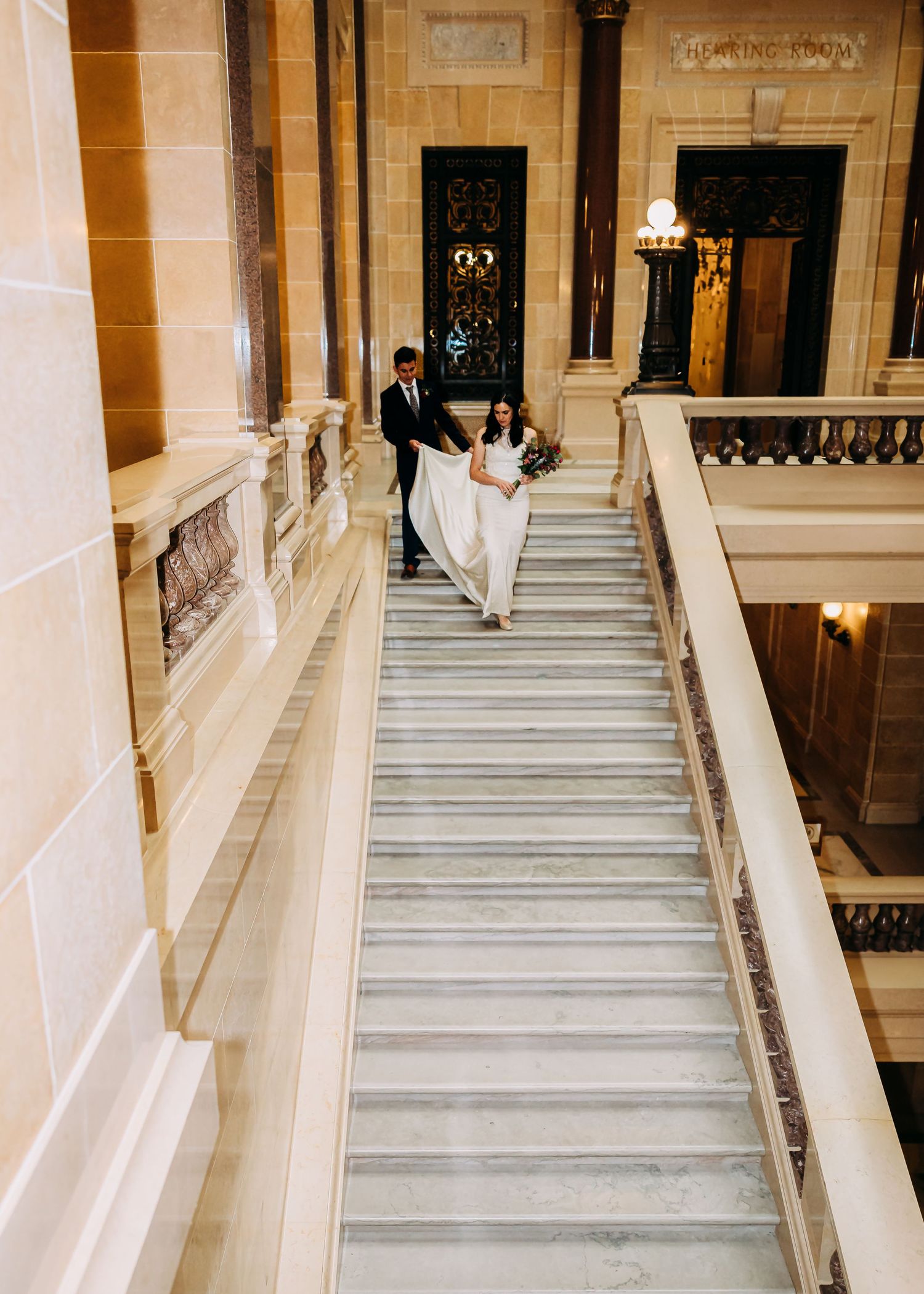 Bride and groom walking down grand marble staircase in elegant building during wedding day photos.