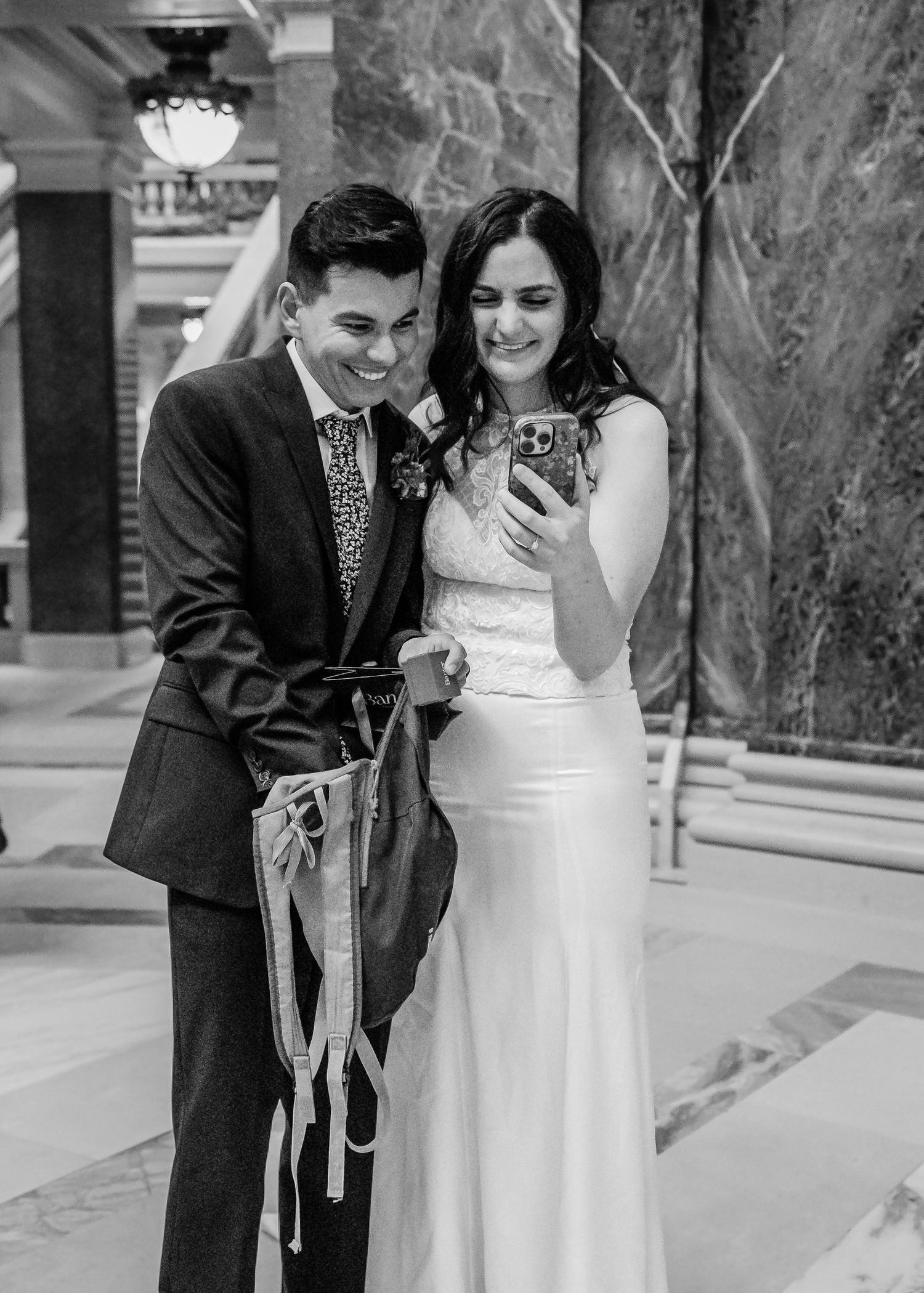 Laughing newlyweds in black and white photo looking at phone, bride holding colorful ribbons.