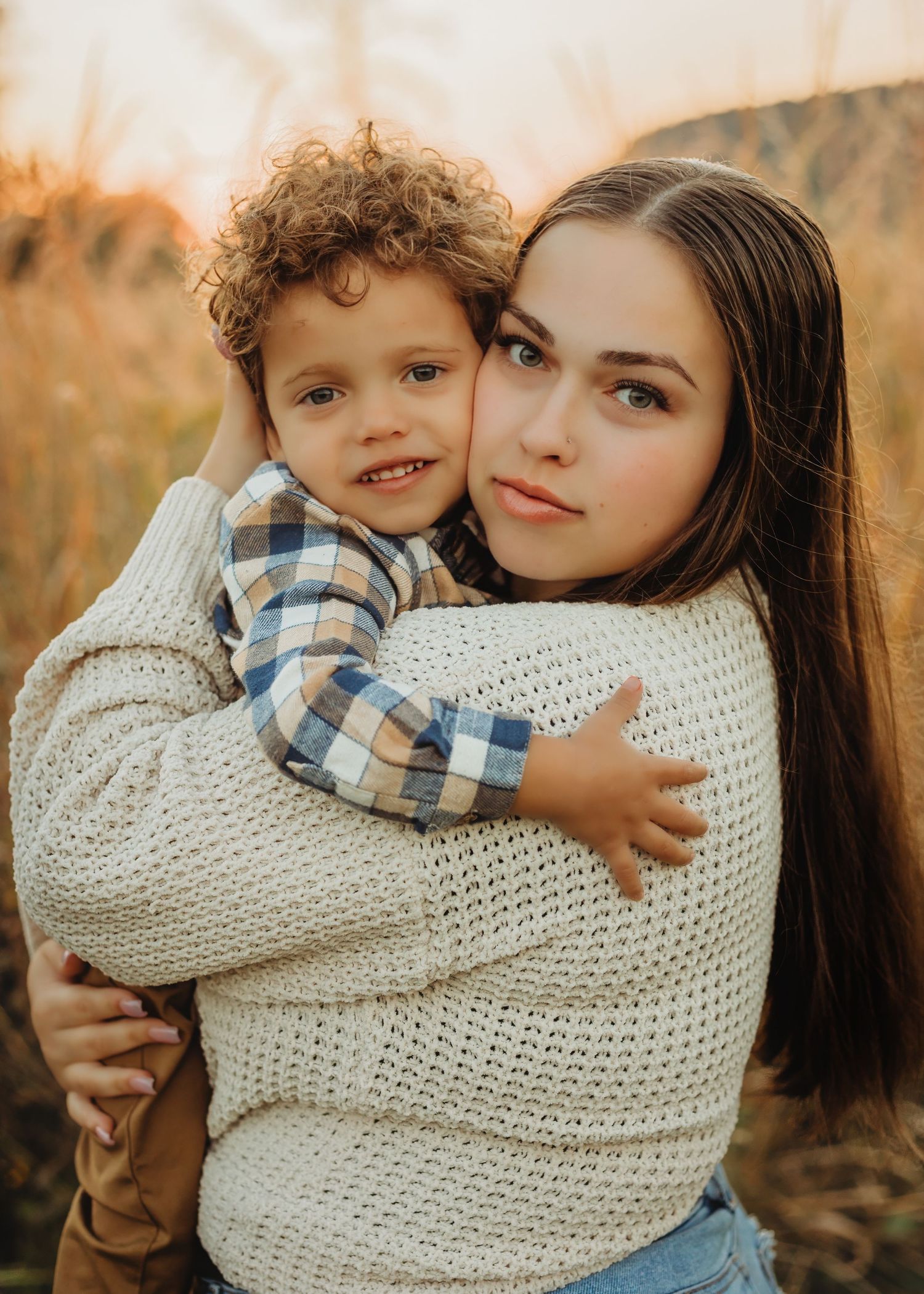 Young brunette woman in cream sweater tenderly embracing curly-haired toddler boy in golden field.