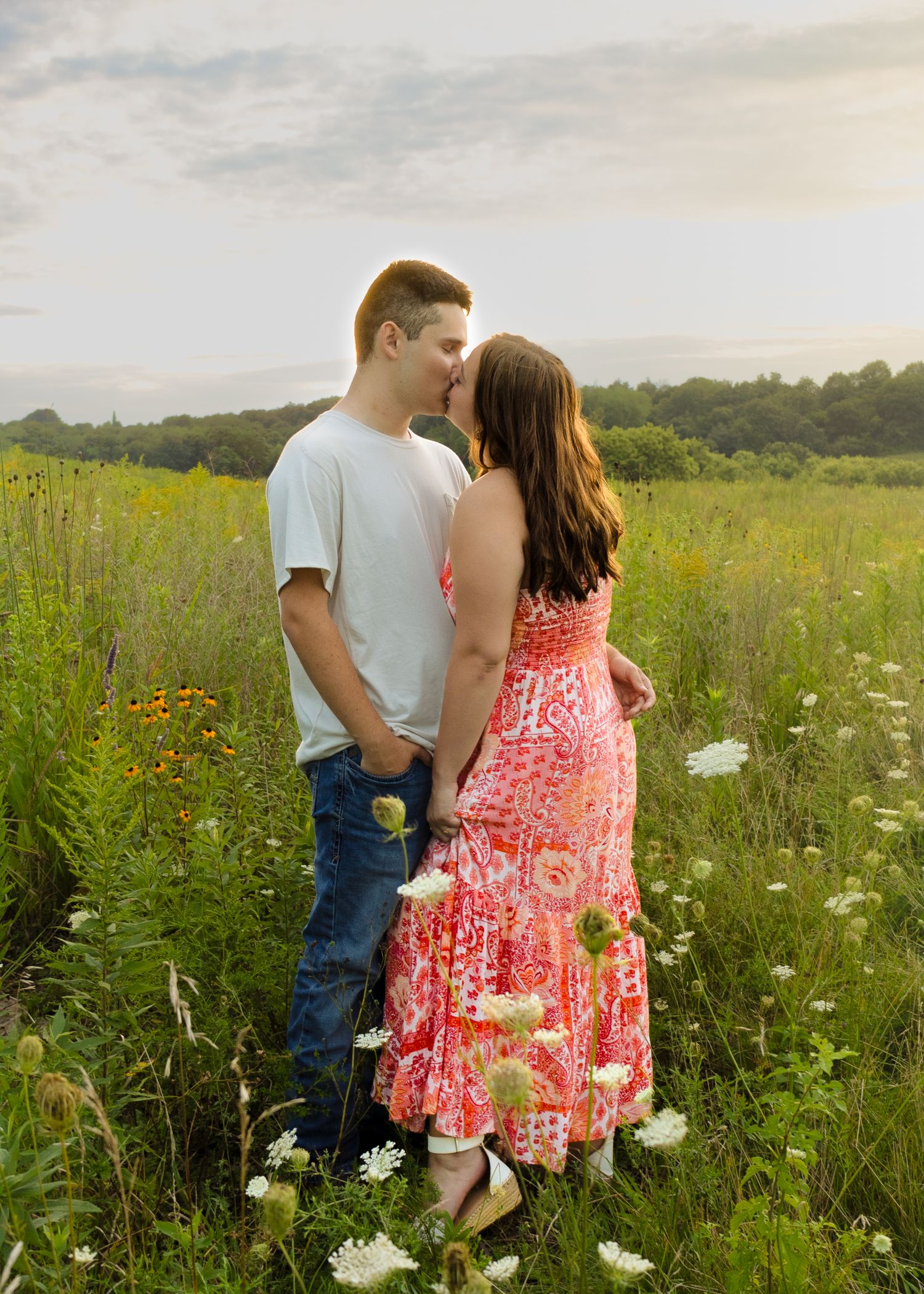 Young couple sharing a kiss in a vibrant wildflower meadow, woman in pink floral maxi dress at sunset.