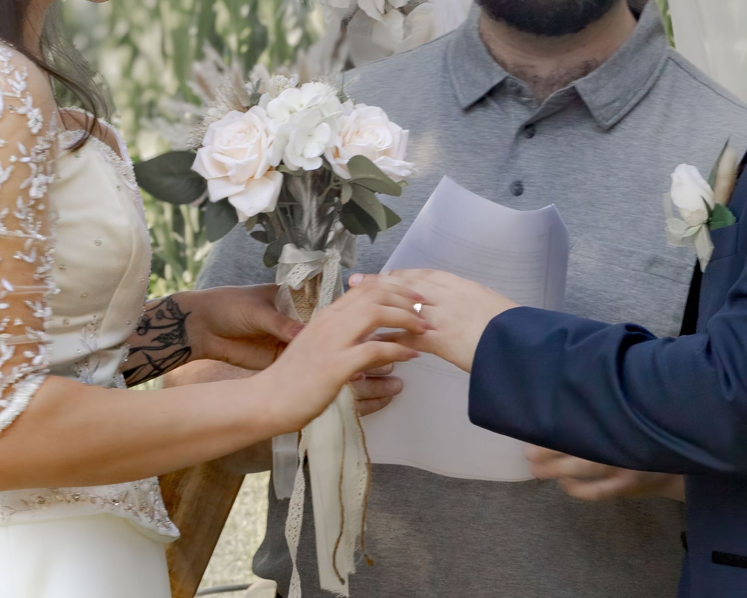 Groom placing a wedding ring on brides finger during outdoor ceremony, bride holding white rose bouquet.