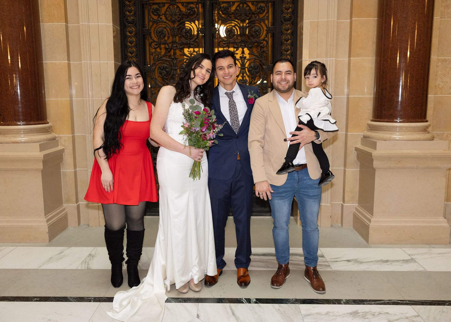 A bride and groom pose with three guests, one holding a toddler, before ornate golden doors.