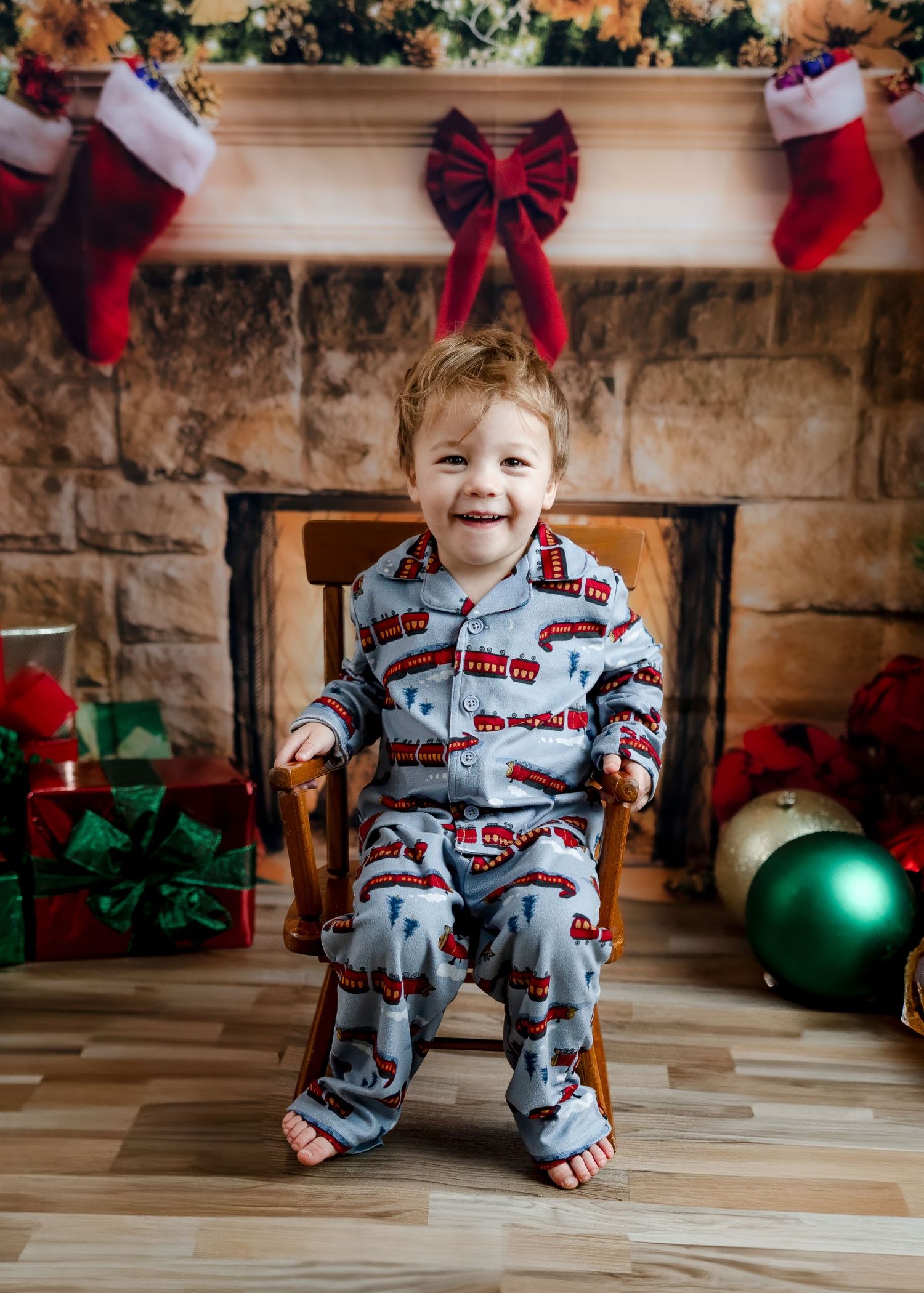 Happy toddler in blue train-print pajamas smiling in rocking chair by decorated Christmas fireplace.