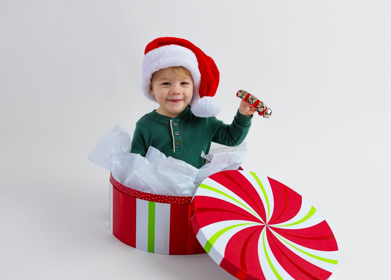 A smiling toddler wearing a Santa hat pops out of a large red and white peppermint gift box holding a candy cane.