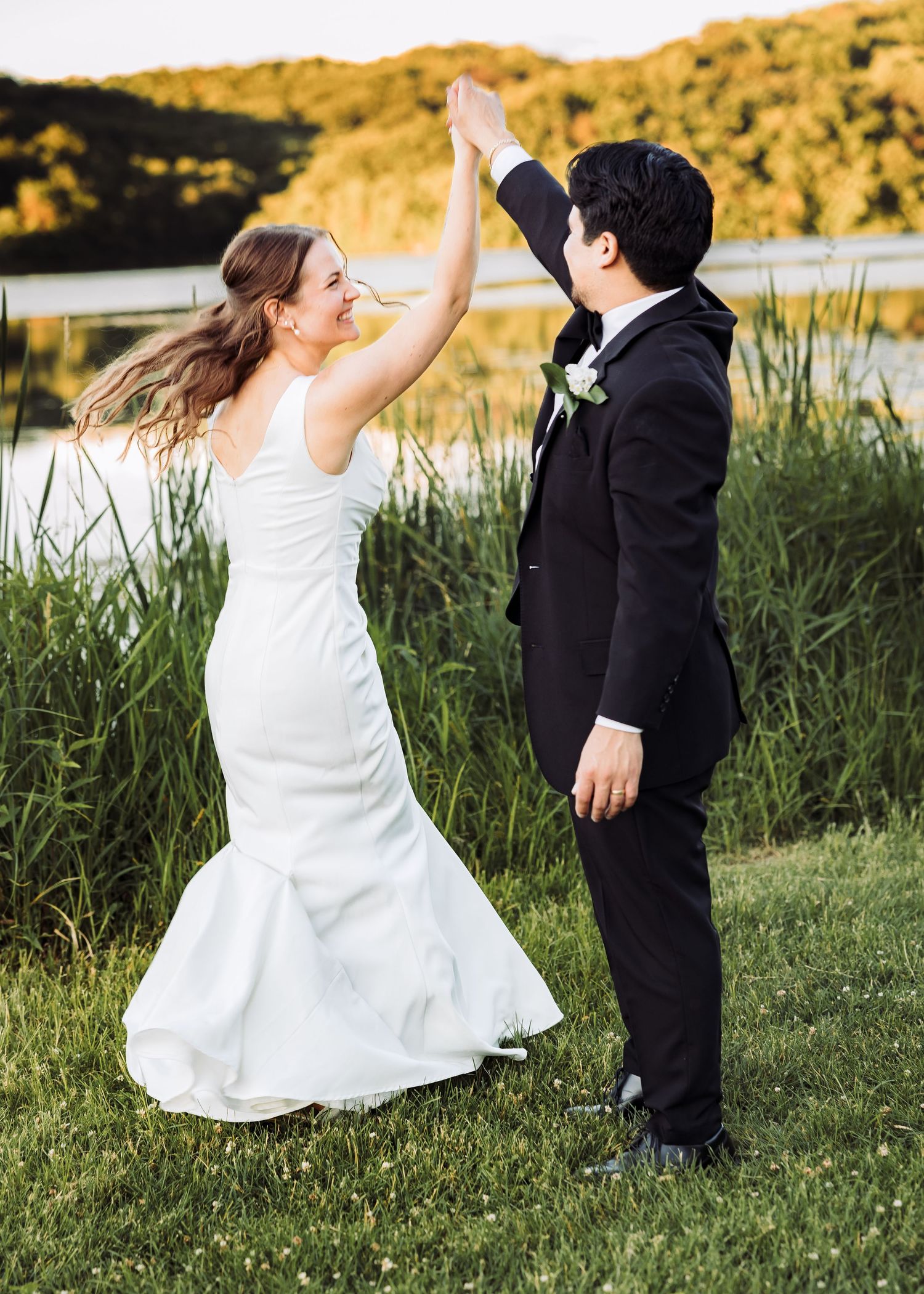Bride and groom high-fiving outdoors near a lake, wearing wedding attire, surrounded by tall green grass.