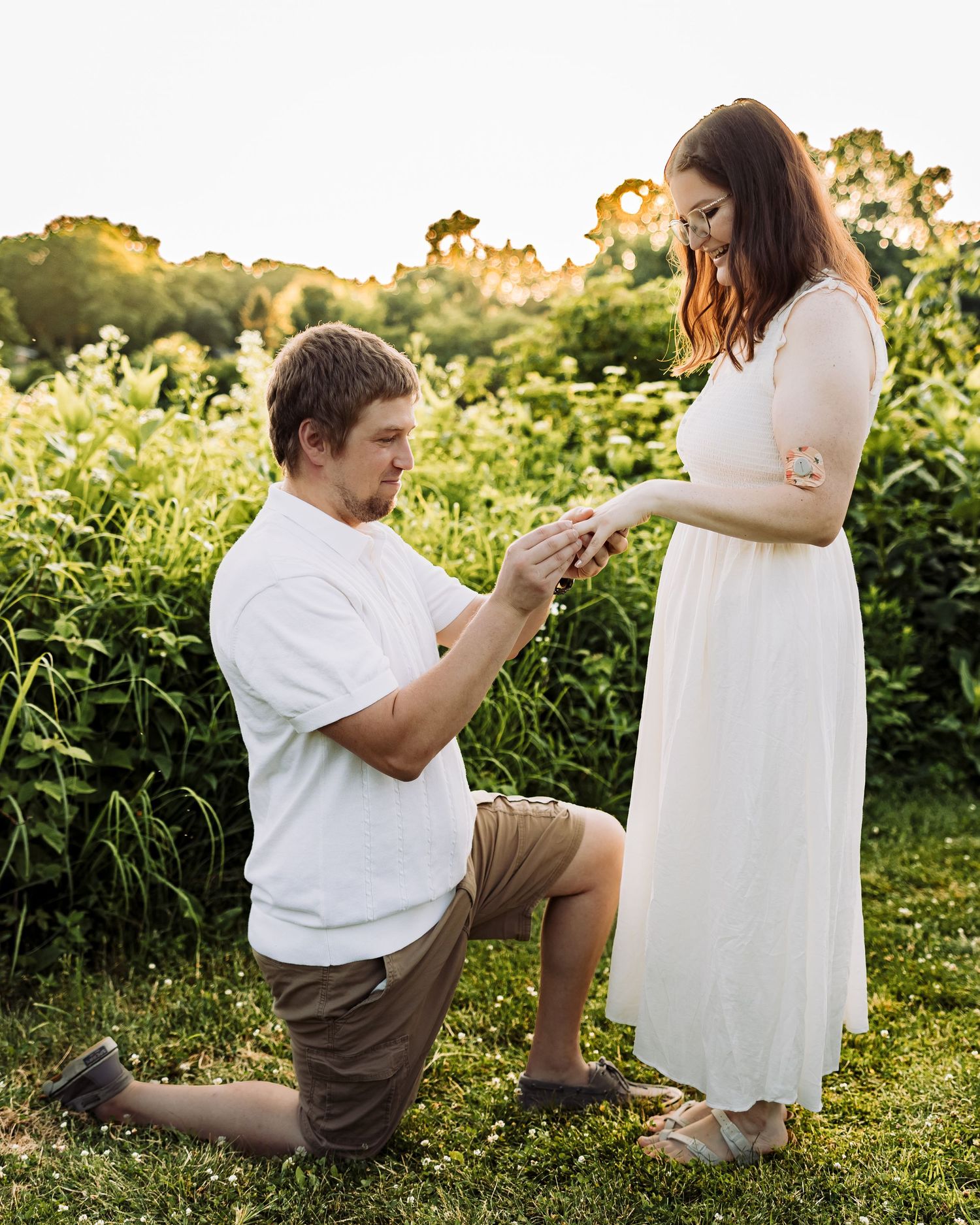 Man kneeling on one knee proposing to woman in white dress outdoors at golden hour in lush green garden.