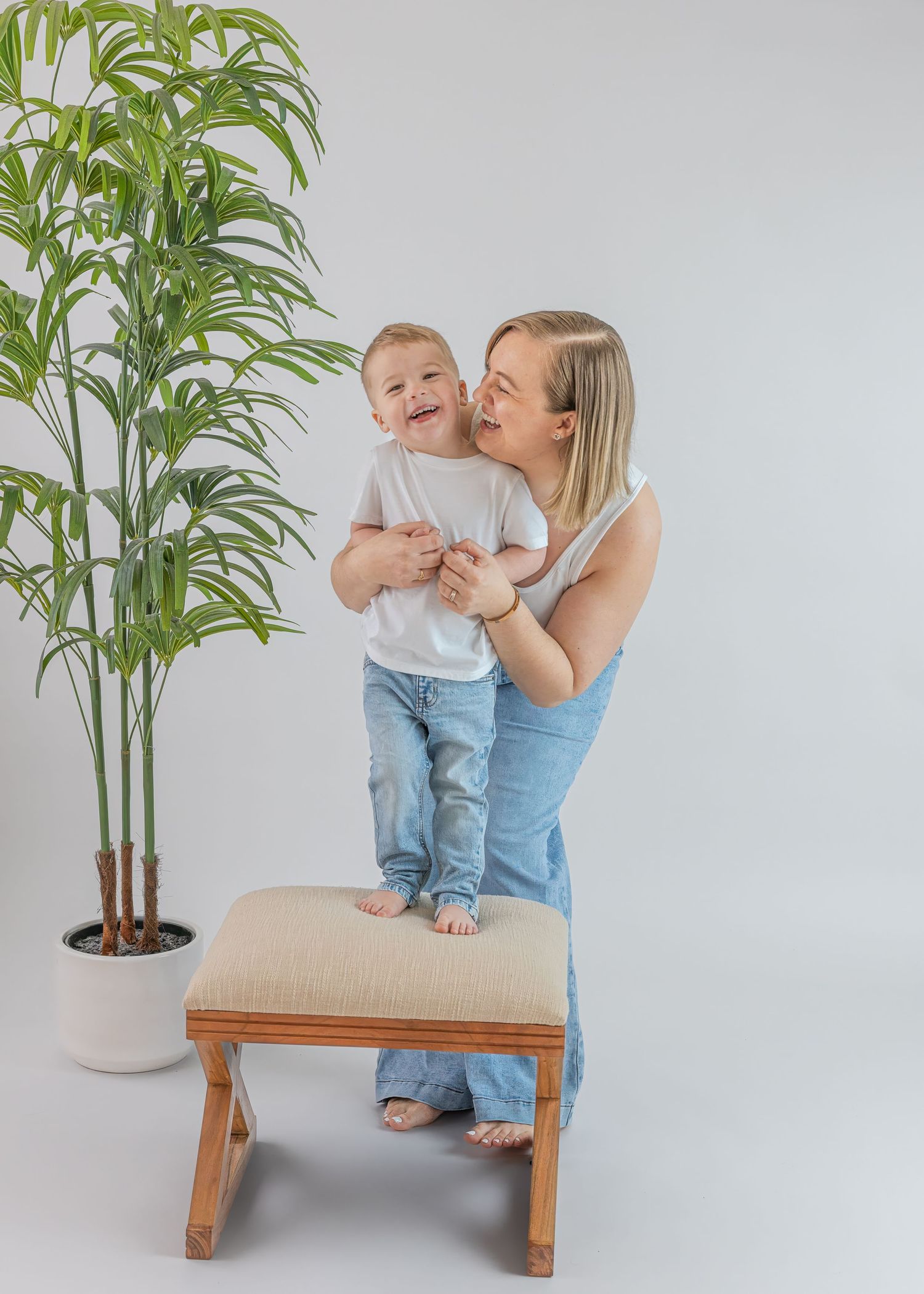 Joyful blonde mother lifting laughing toddler son standing on bench beside potted palm plant.