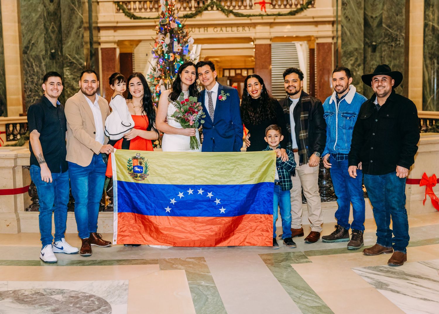Venezuelan wedding party poses with flag near Christmas tree in elegant marble hall.