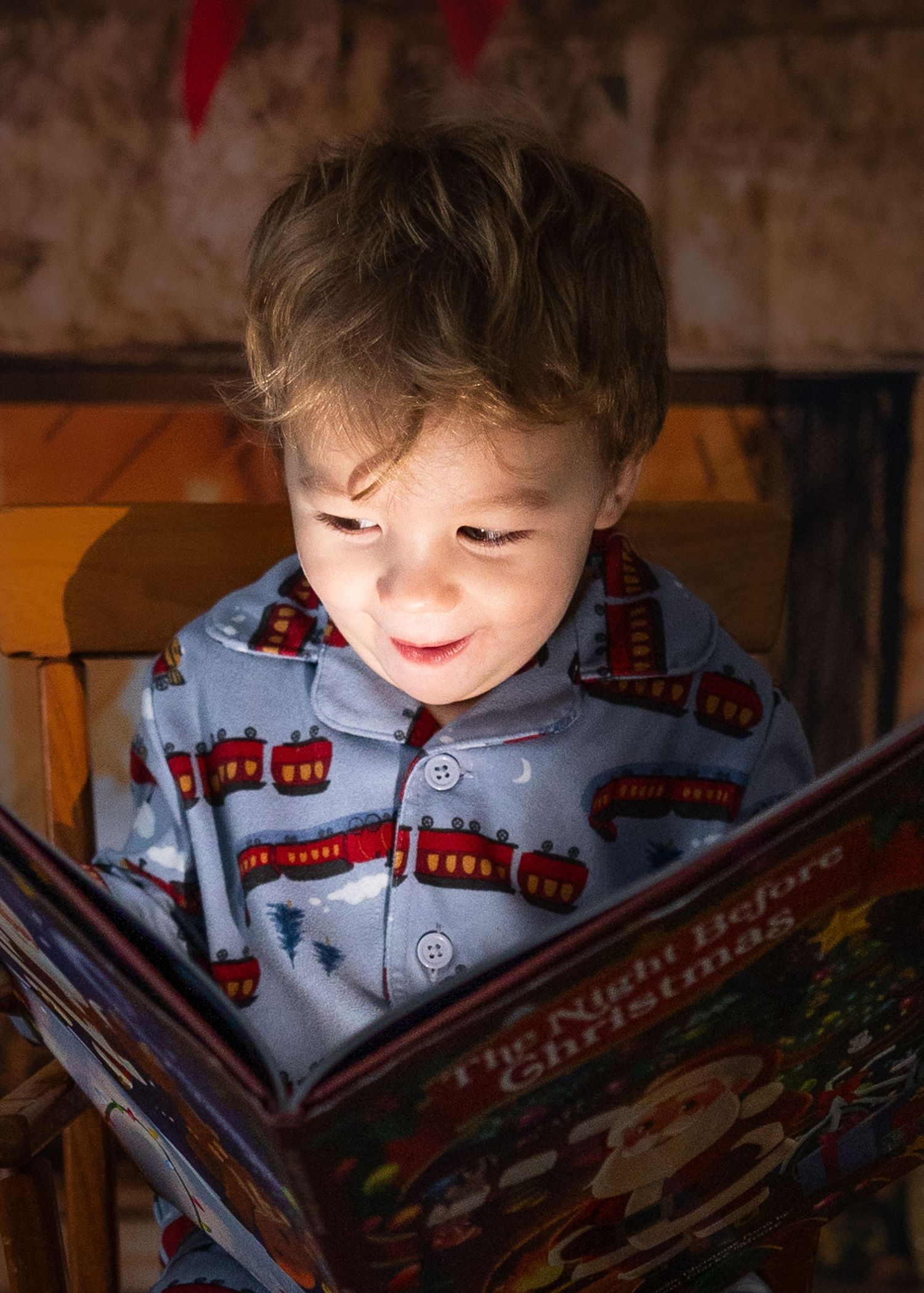Smiling toddler in blue train-print pajamas reading The Night Before Christmas book.