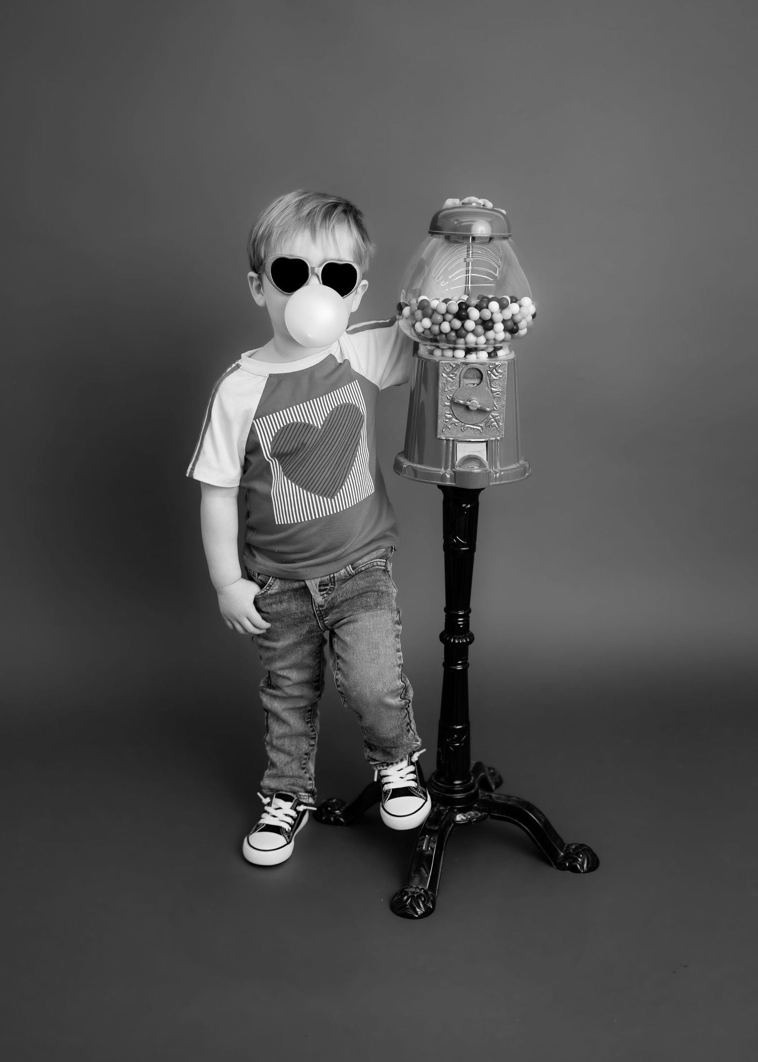 Black and white photo of cool toddler in heart sunglasses posing by vintage gumball machine.