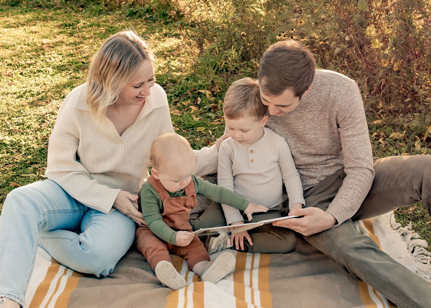 A loving family of four reads a book together on a blanket in a warm golden autumn outdoor setting.