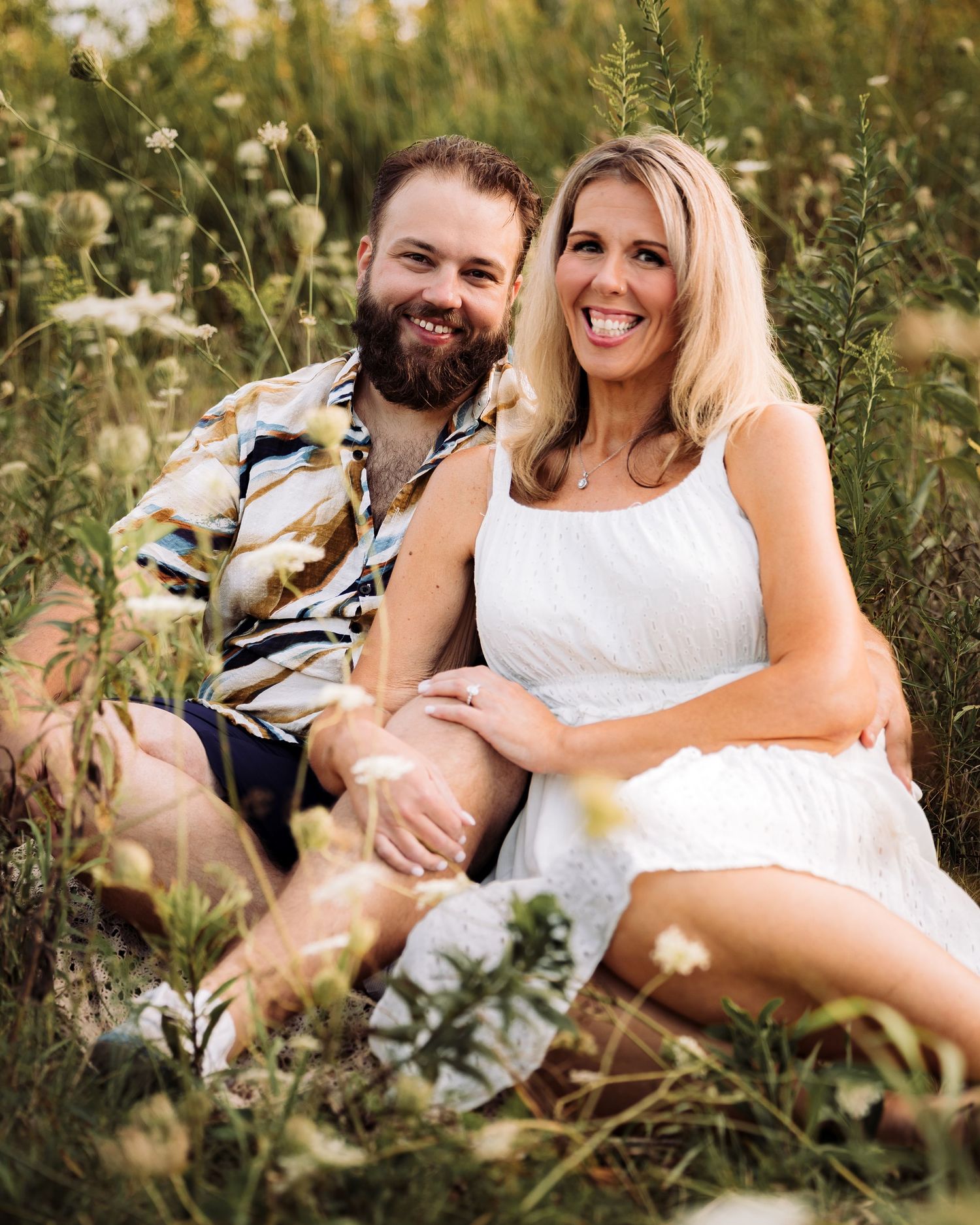 A smiling couple sits together in a wildflower meadow, woman in white dress, man in patterned shirt.