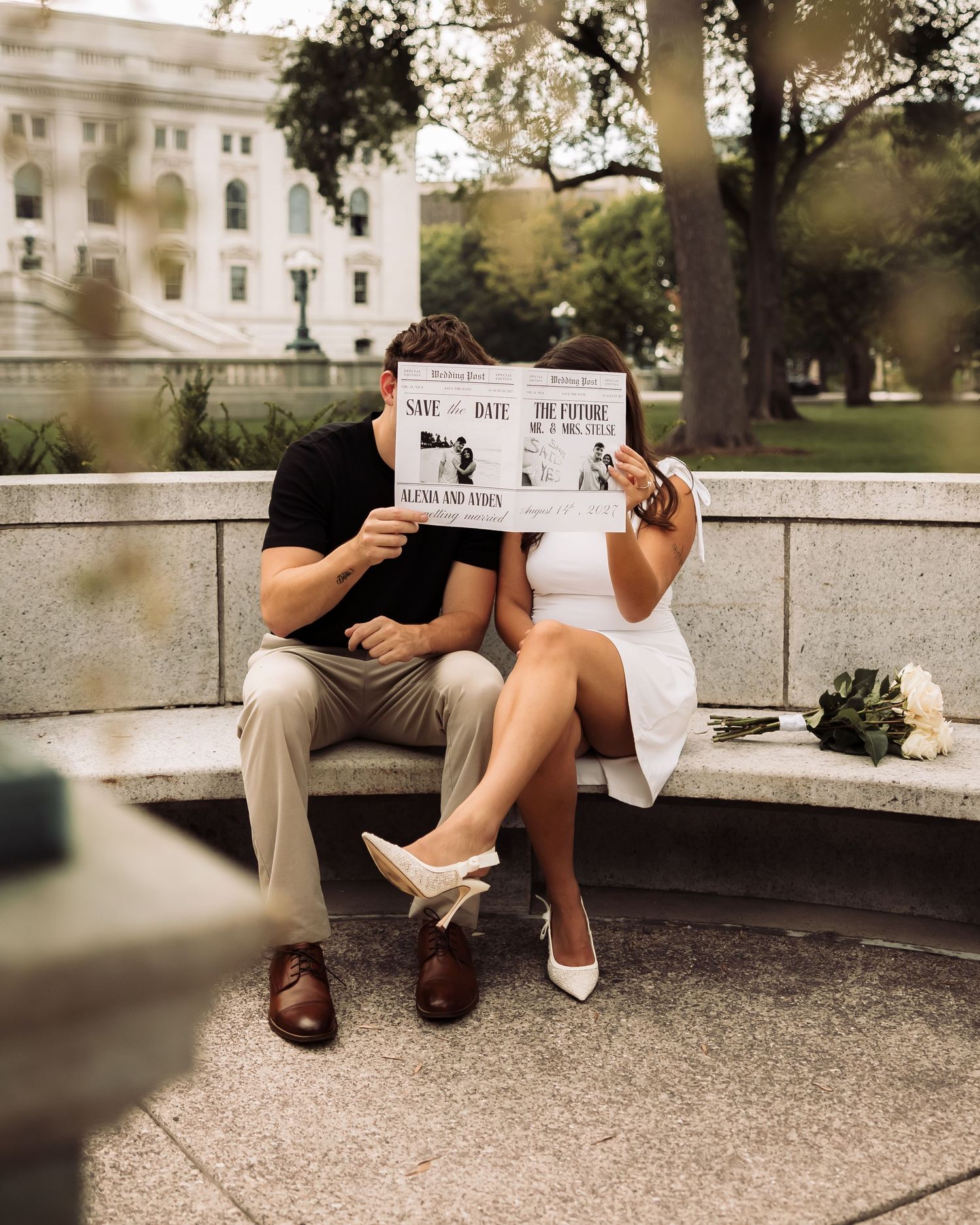 Couple sitting on stone bench holding save the date newspaper in front of faces, white dress, bouquet nearby, classic building backdrop.