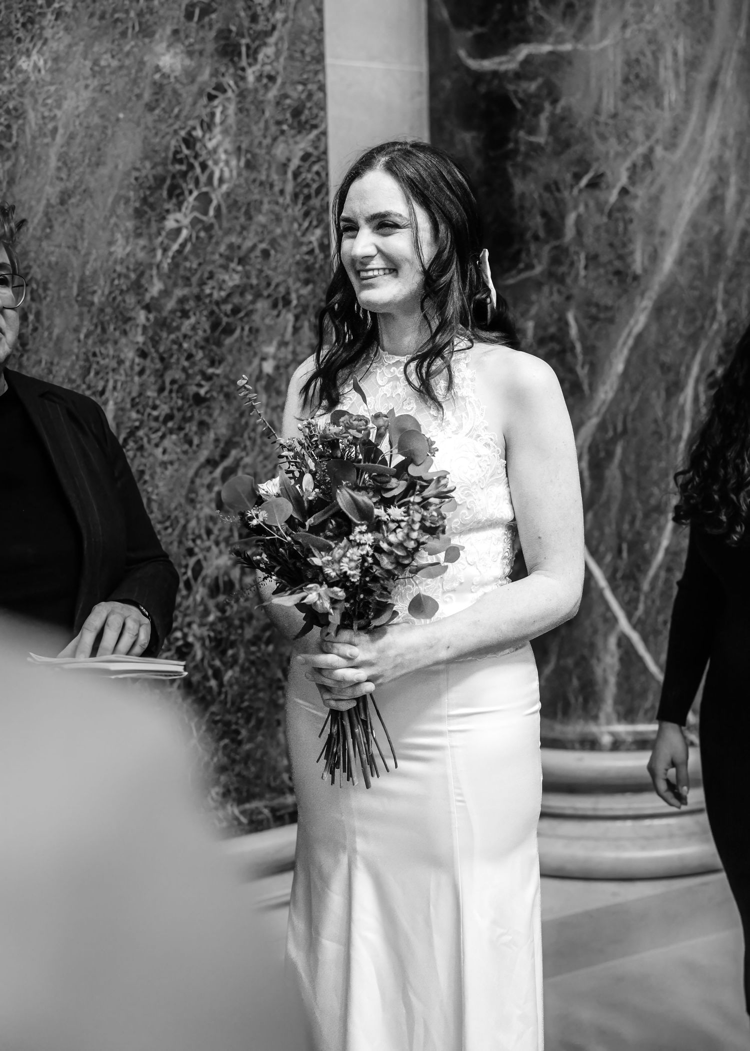 Radiant bride in lace white gown smiles while holding wildflower bouquet during wedding ceremony.
