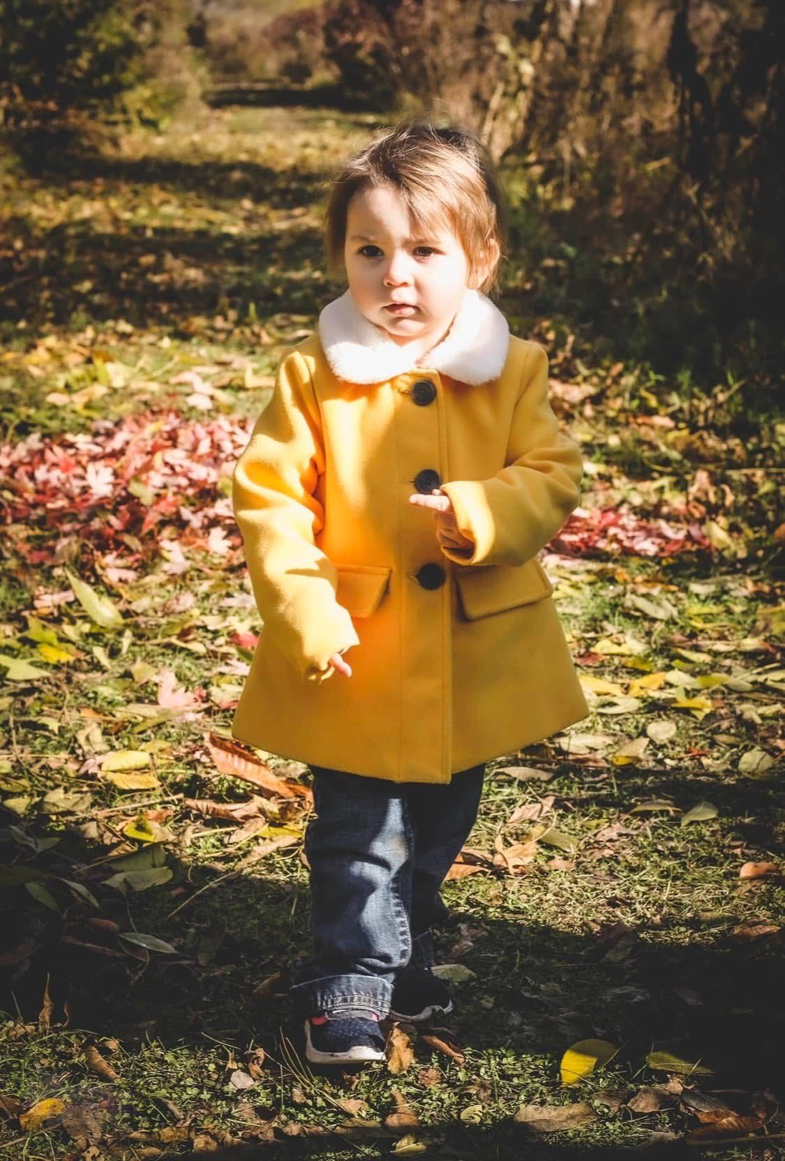 Toddler in yellow coat with white collar walks through colorful fallen autumn leaves outdoors.