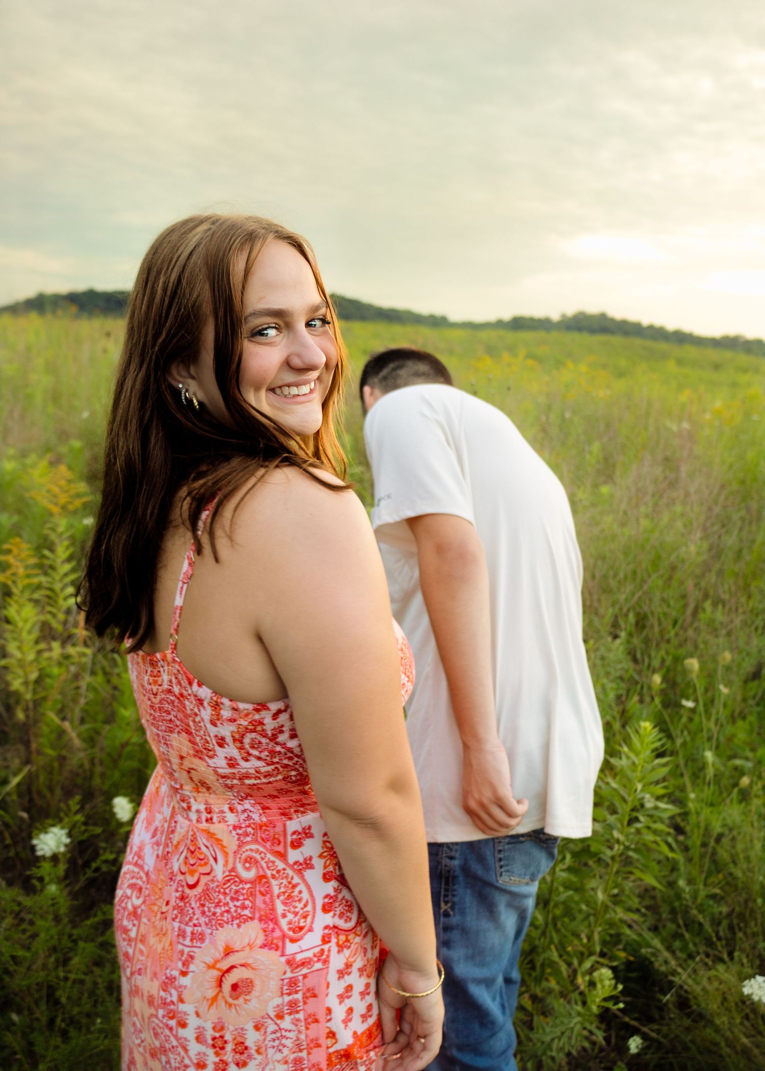 Smiling young woman in pink floral dress looking back at camera while man bends down in green meadow.