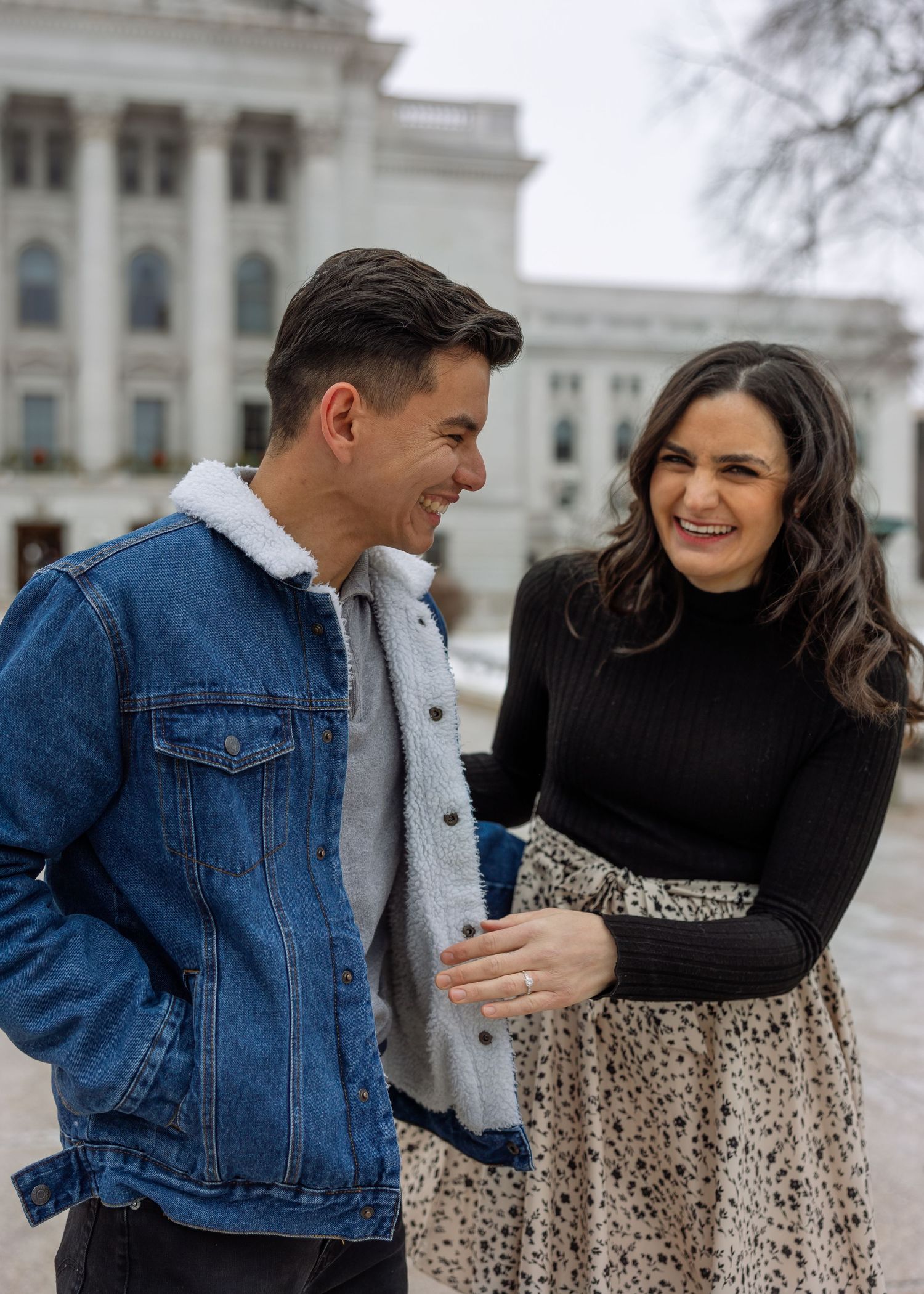A laughing couple walks arm in arm outside a historic domed capitol building on a cloudy winter day.