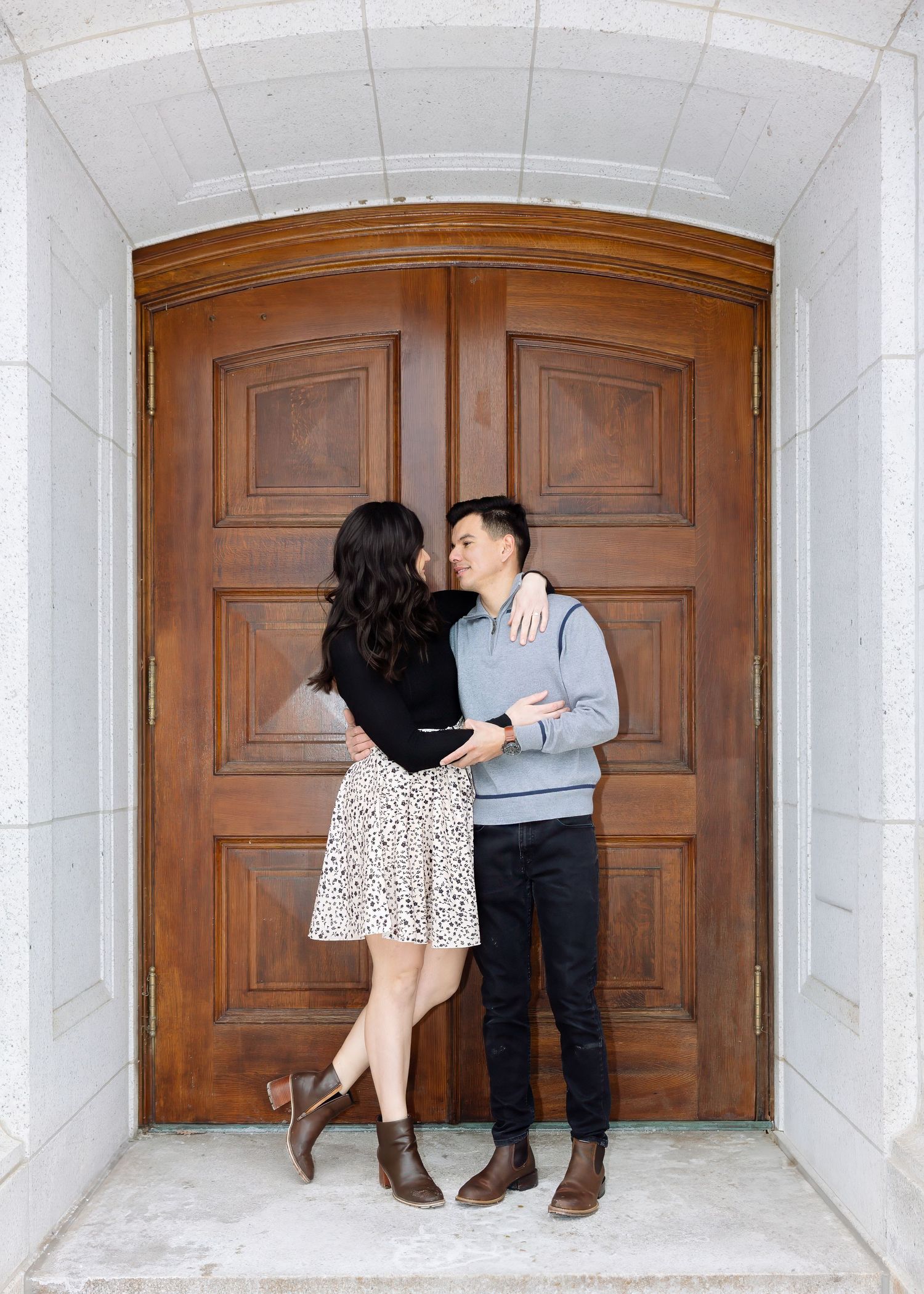 A couple gazes at each other romantically in front of grand wooden arched doors at a stone building.
