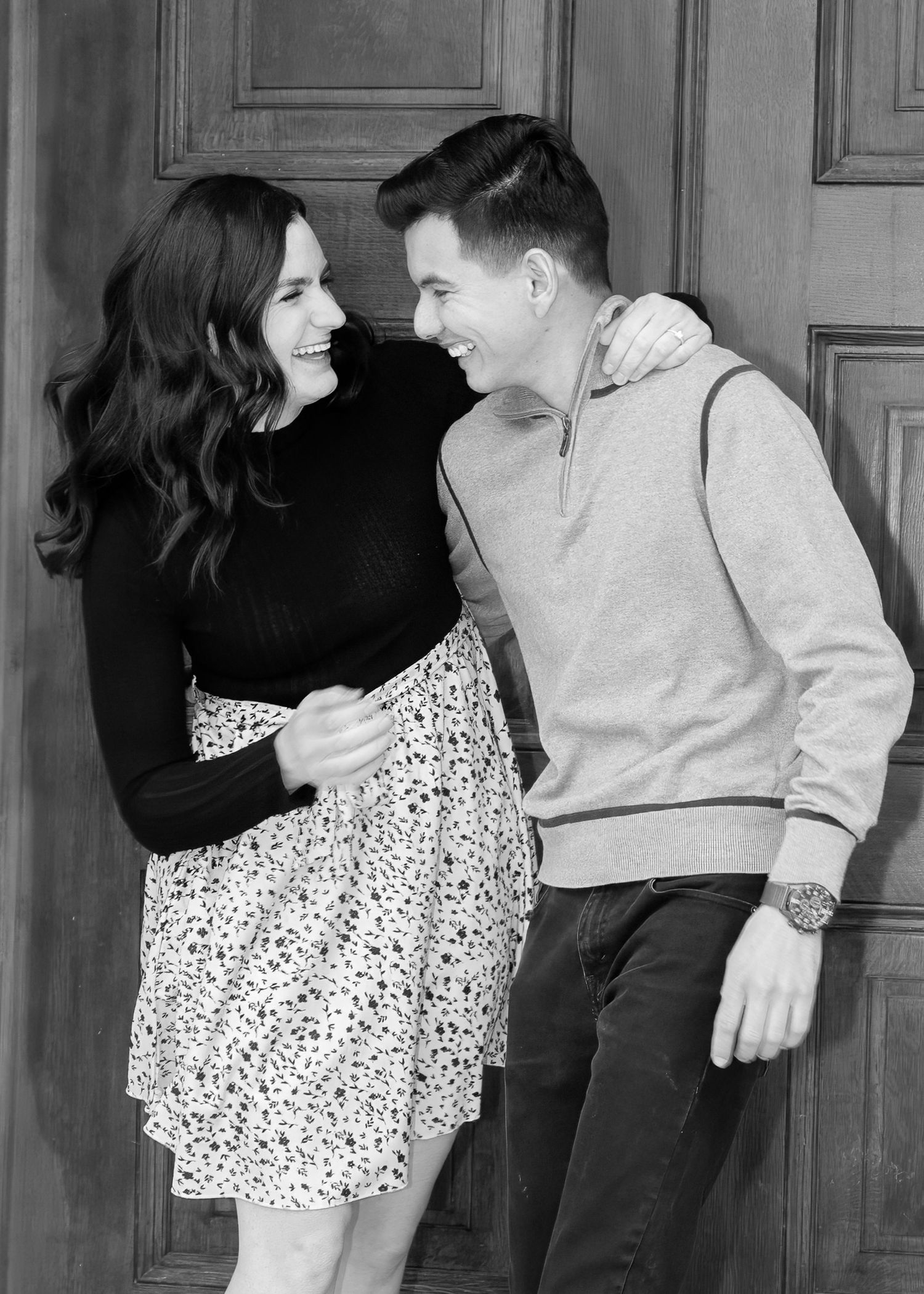 Black and white photo of a laughing couple holding hands in front of a wooden door.