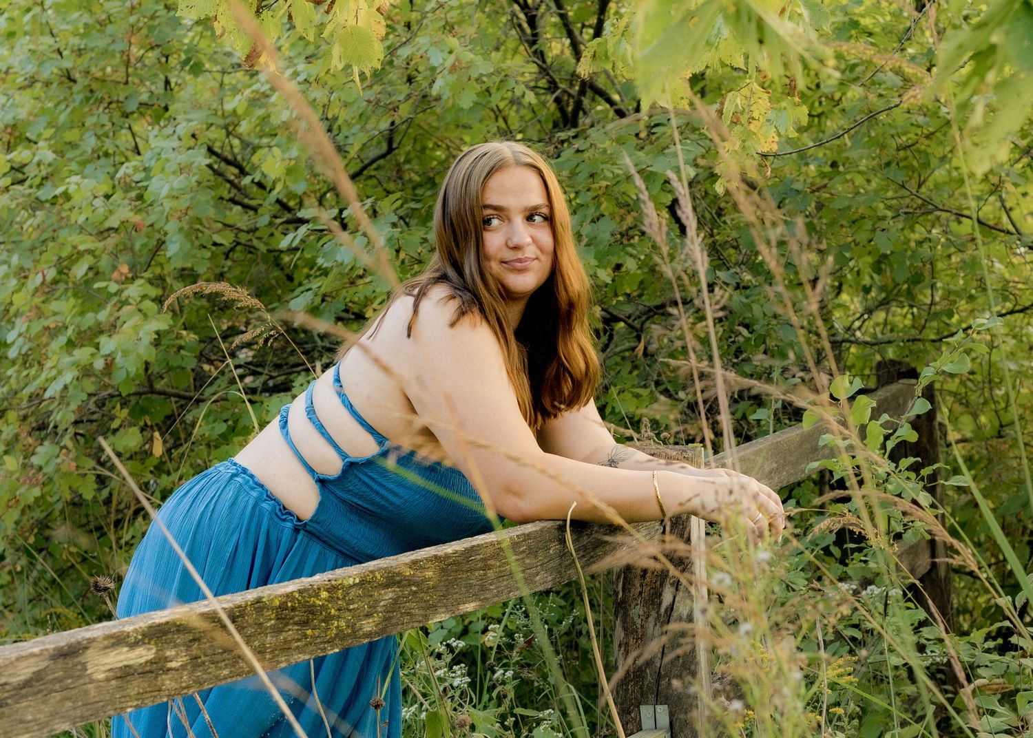 Senior girl in teal blue dress leans on rustic wooden fence surrounded by lush green foliage.