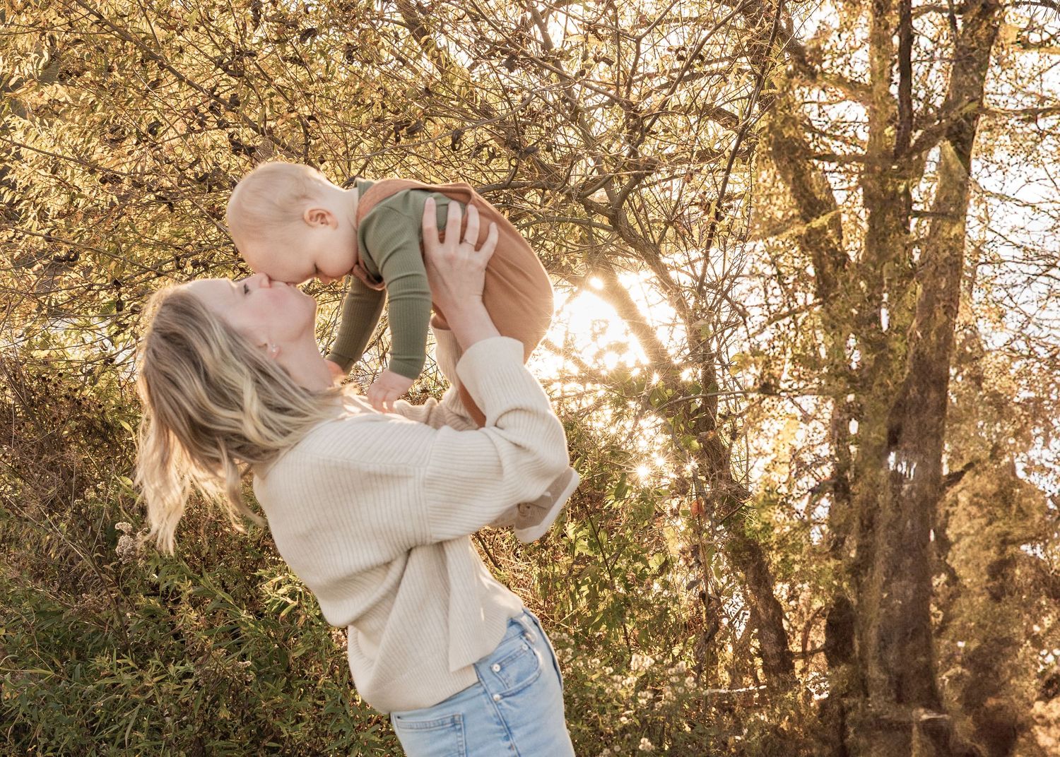 A blonde mother lifts her smiling baby up in a sunlit outdoor autumn setting.