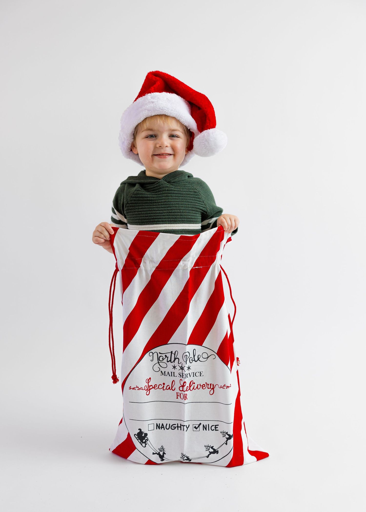 Smiling child in green sweater and Santa hat holds open a red striped North Pole delivery sack.