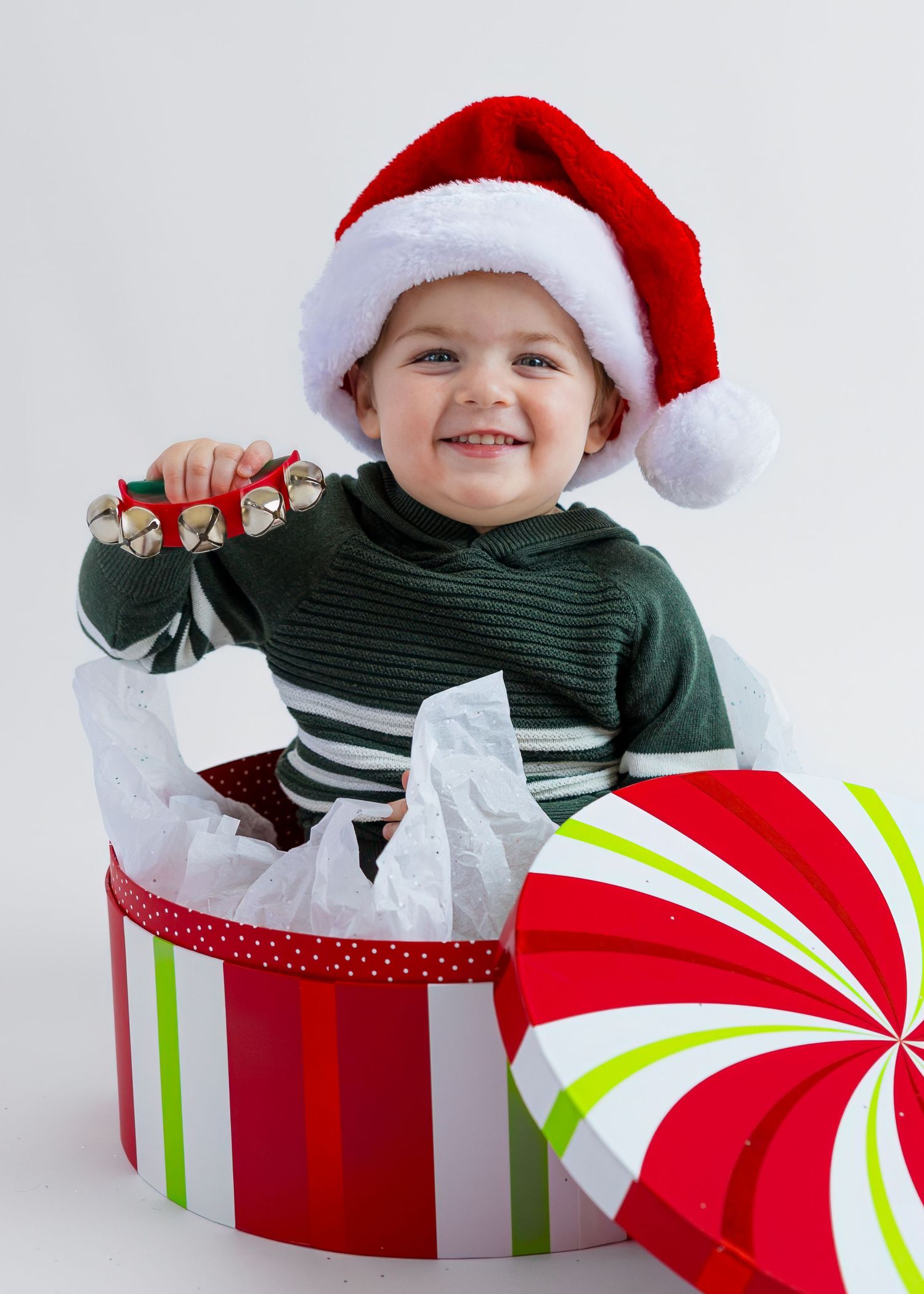 Happy toddler in Santa hat sitting in festive gift box holding jingle bells for Christmas photo.