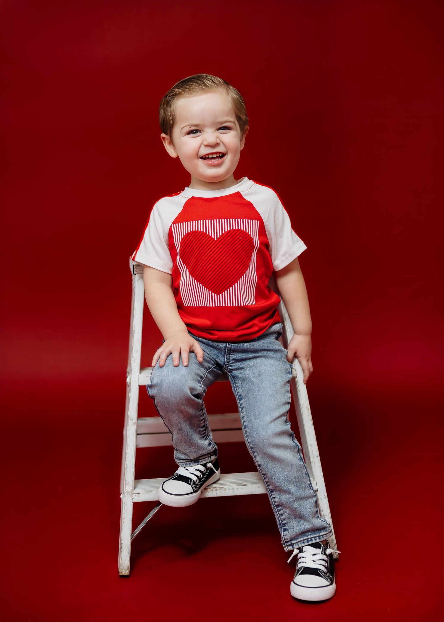 Happy toddler boy in red heart Valentine's Day shirt and jeans sitting on white ladder stool.