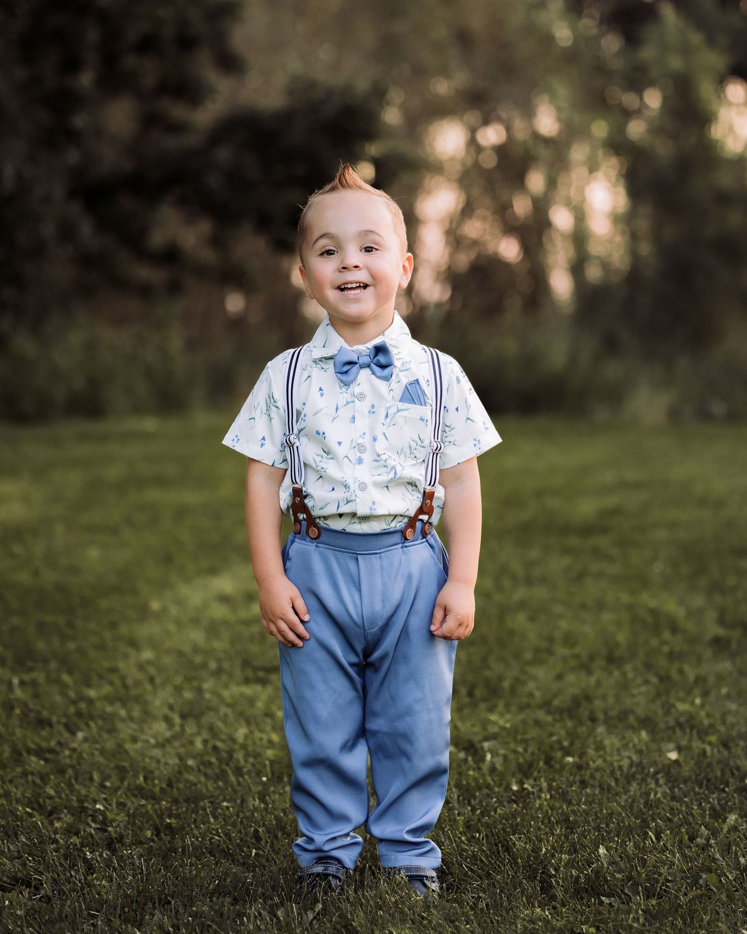 Smiling toddler boy in blue suspenders, bow tie, and dress shirt standing on green grass outdoors.