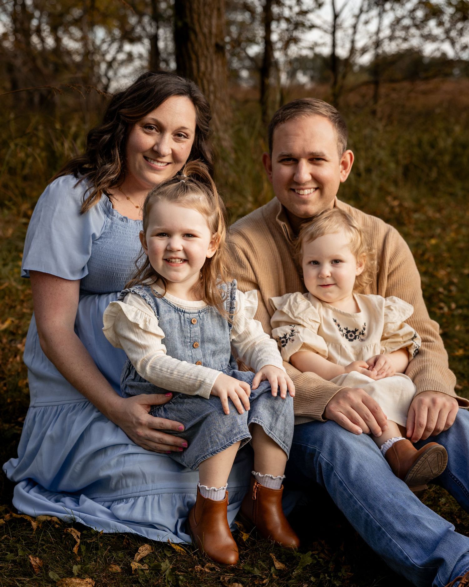 A smiling family of four poses outdoors in autumn, with two young girls sitting on their parents laps wearing coordinated outfits.