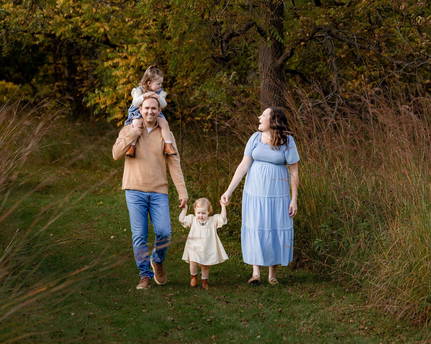 Family of four walking in autumn park, father carrying toddler on shoulders, mother in blue dress holding hands with young child.