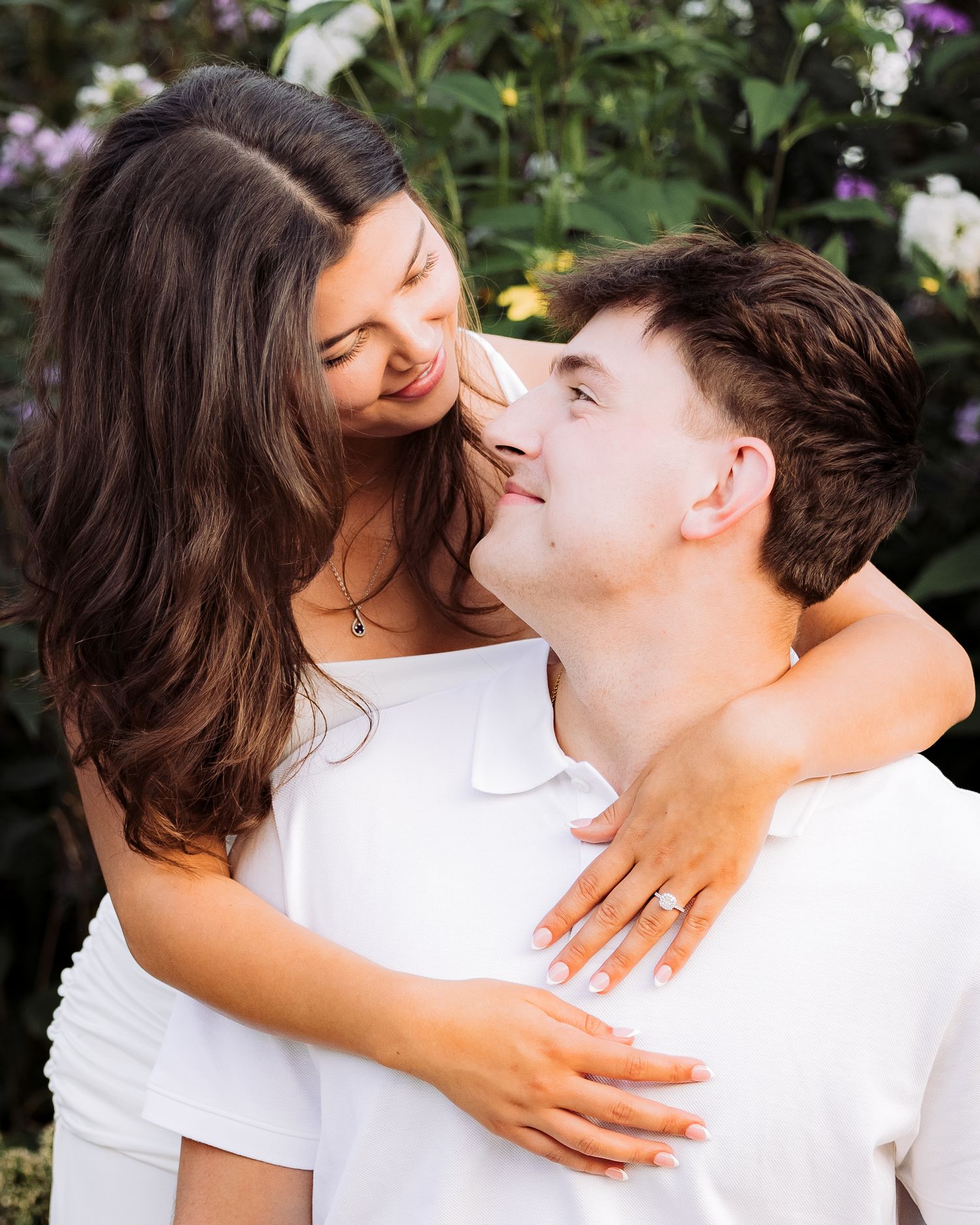 A young couple in white outfits share a romantic moment outdoors, surrounded by colorful blooming flowers.