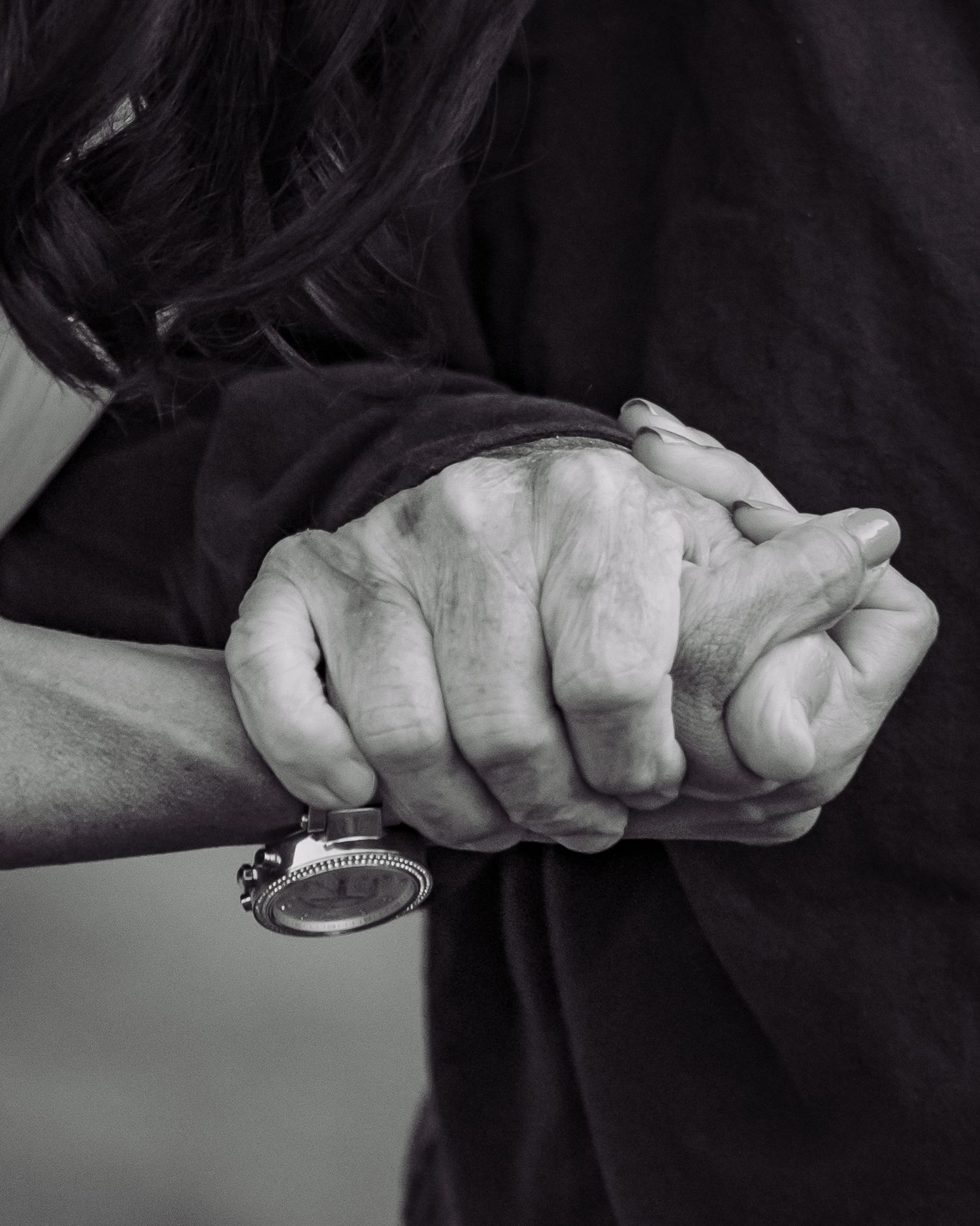 Black and white close-up of two people holding hands, one wearing a watch, against a dark clothing background.