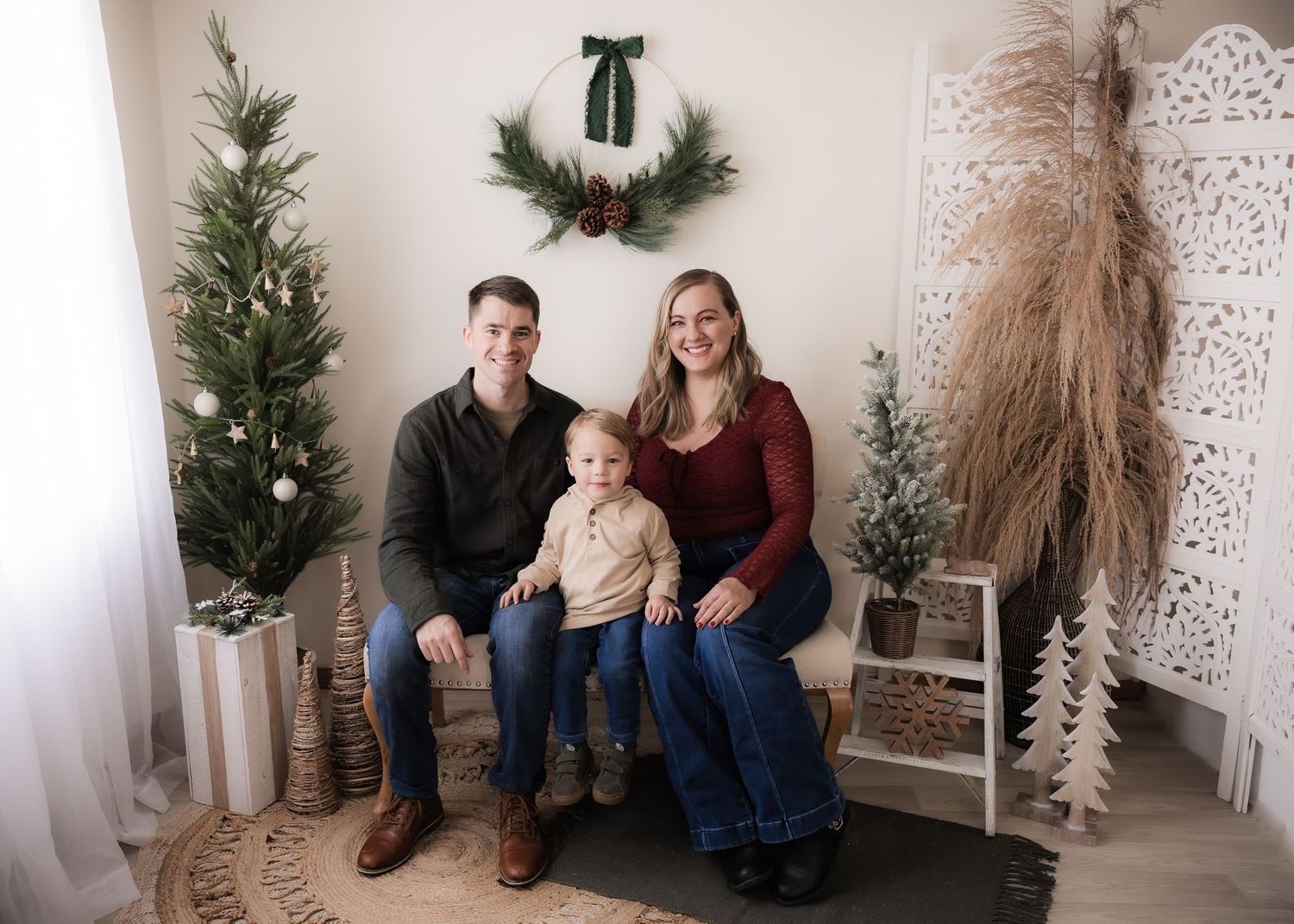 Family of three posing for a Christmas portrait with holiday trees and wreath decor in a cozy studio setting.