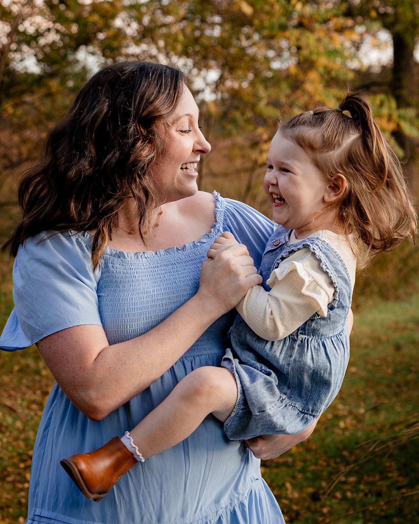 A mother in a blue dress joyfully holds her smiling toddler daughter outdoors among autumn foliage.