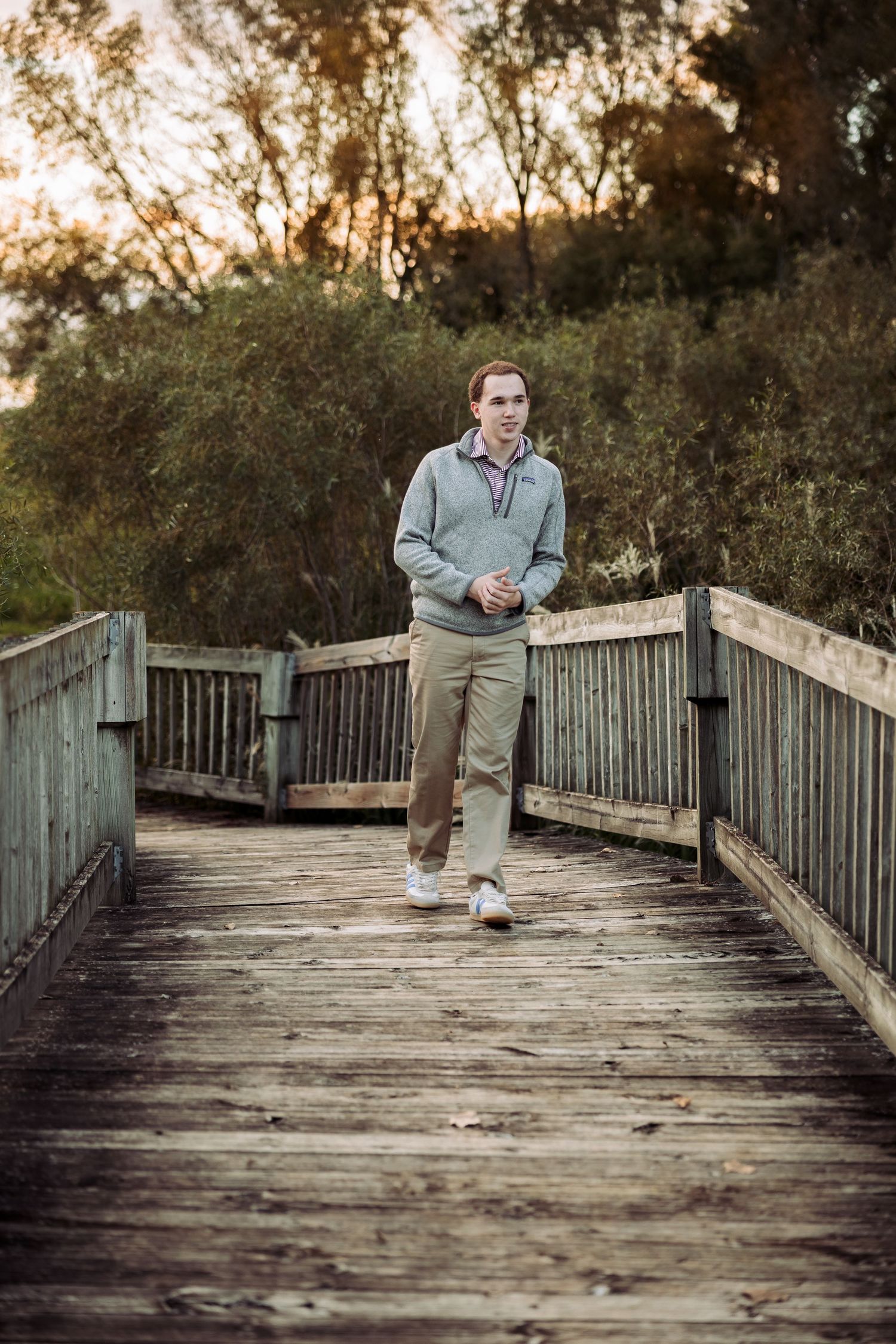 Young man in gray sweater and khaki pants walking on a wooden boardwalk surrounded by lush green trees.