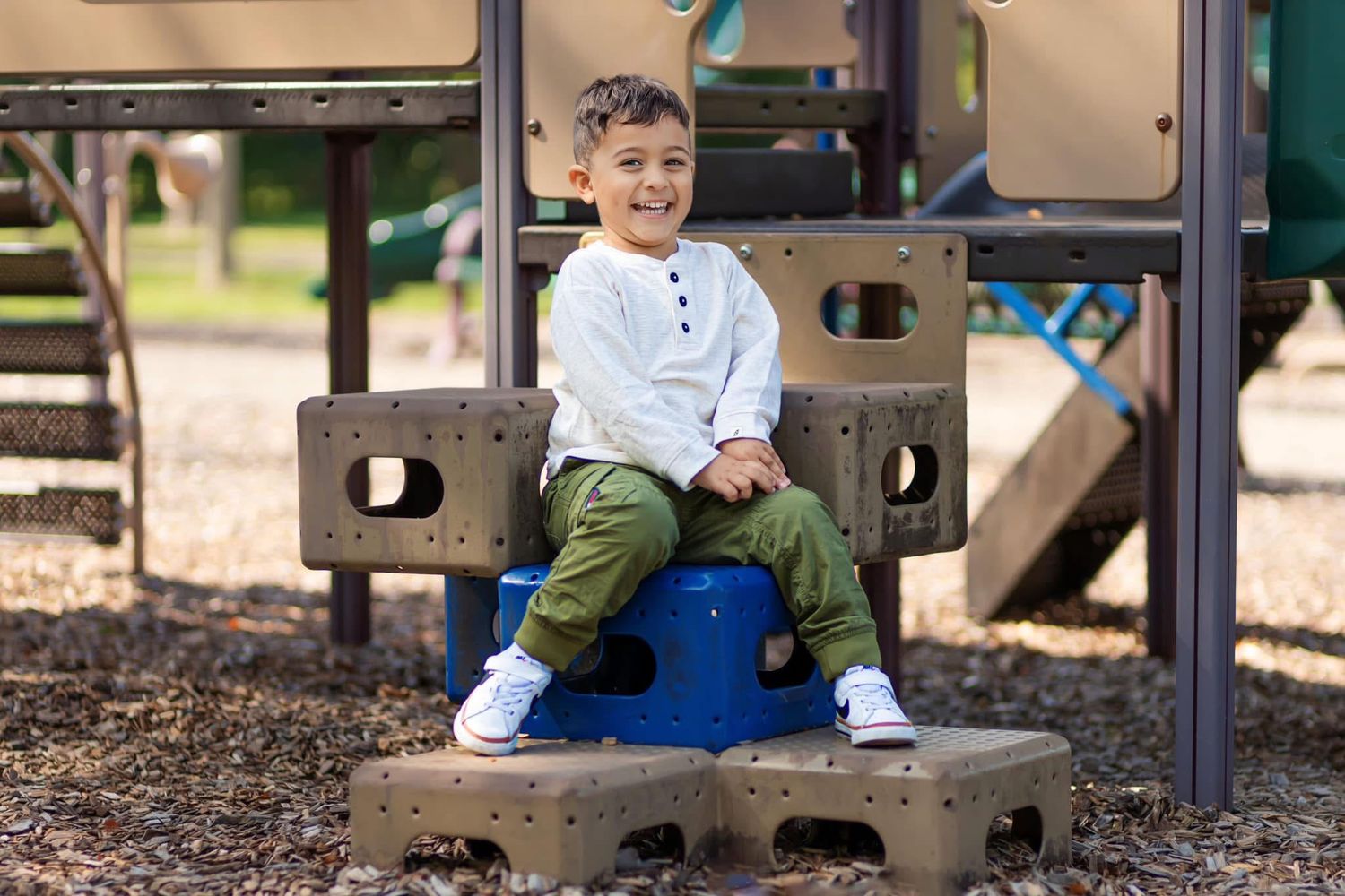 Happy young boy in white shirt and green pants sits smiling at outdoor playground.