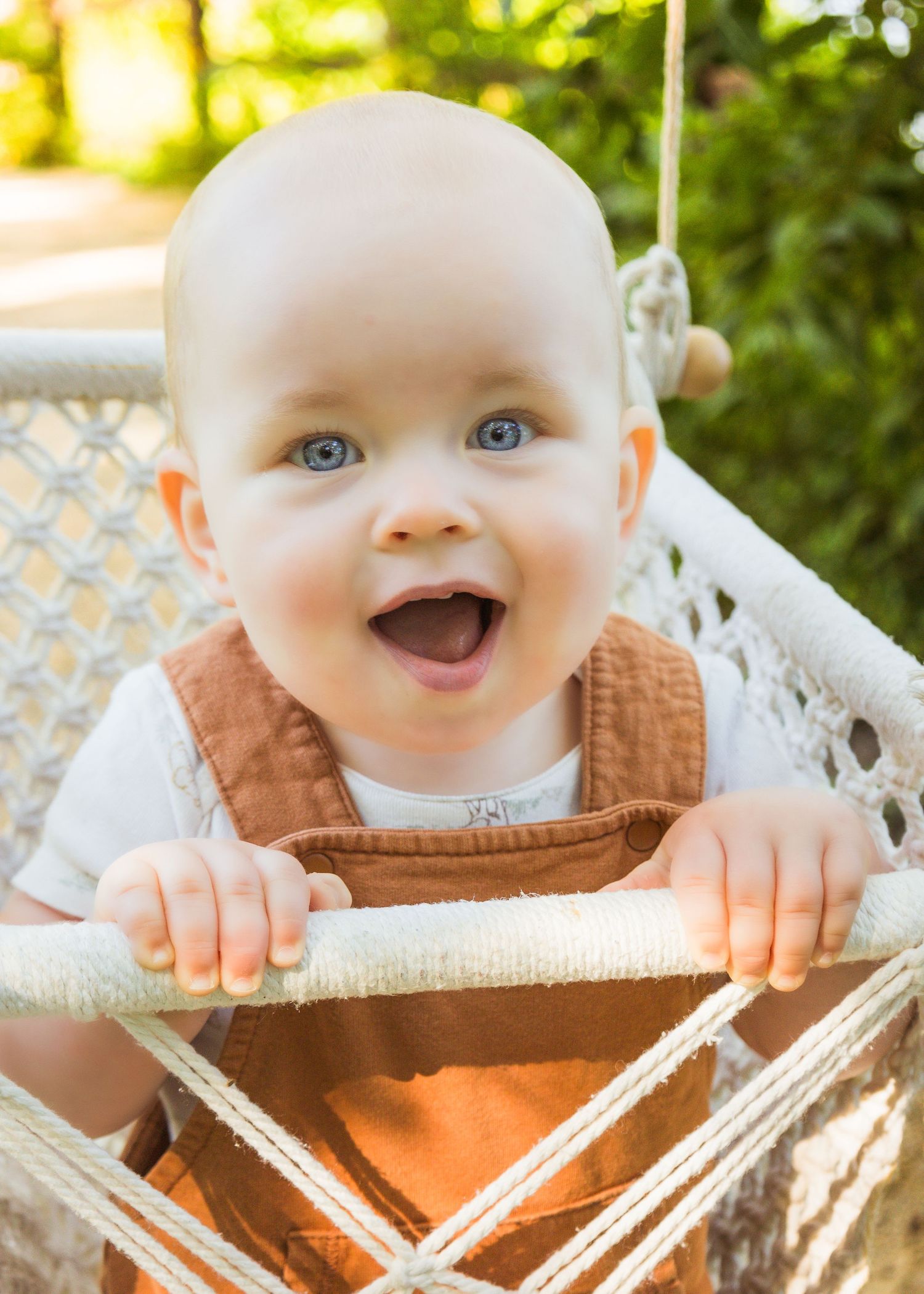 Laughing blue-eyed baby in brown overalls sits in hanging rope swing outdoors.
