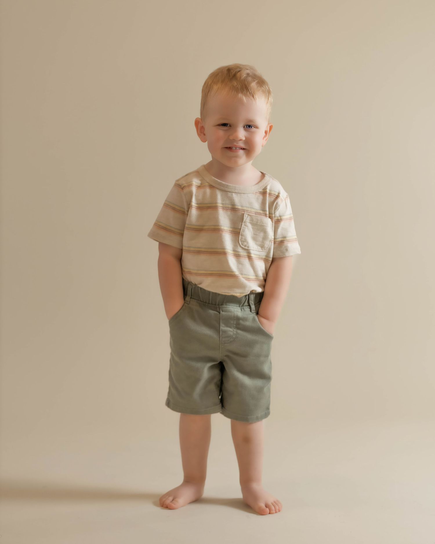 Toddler boy in striped tee and sage green shorts stands barefoot smiling on a neutral background.